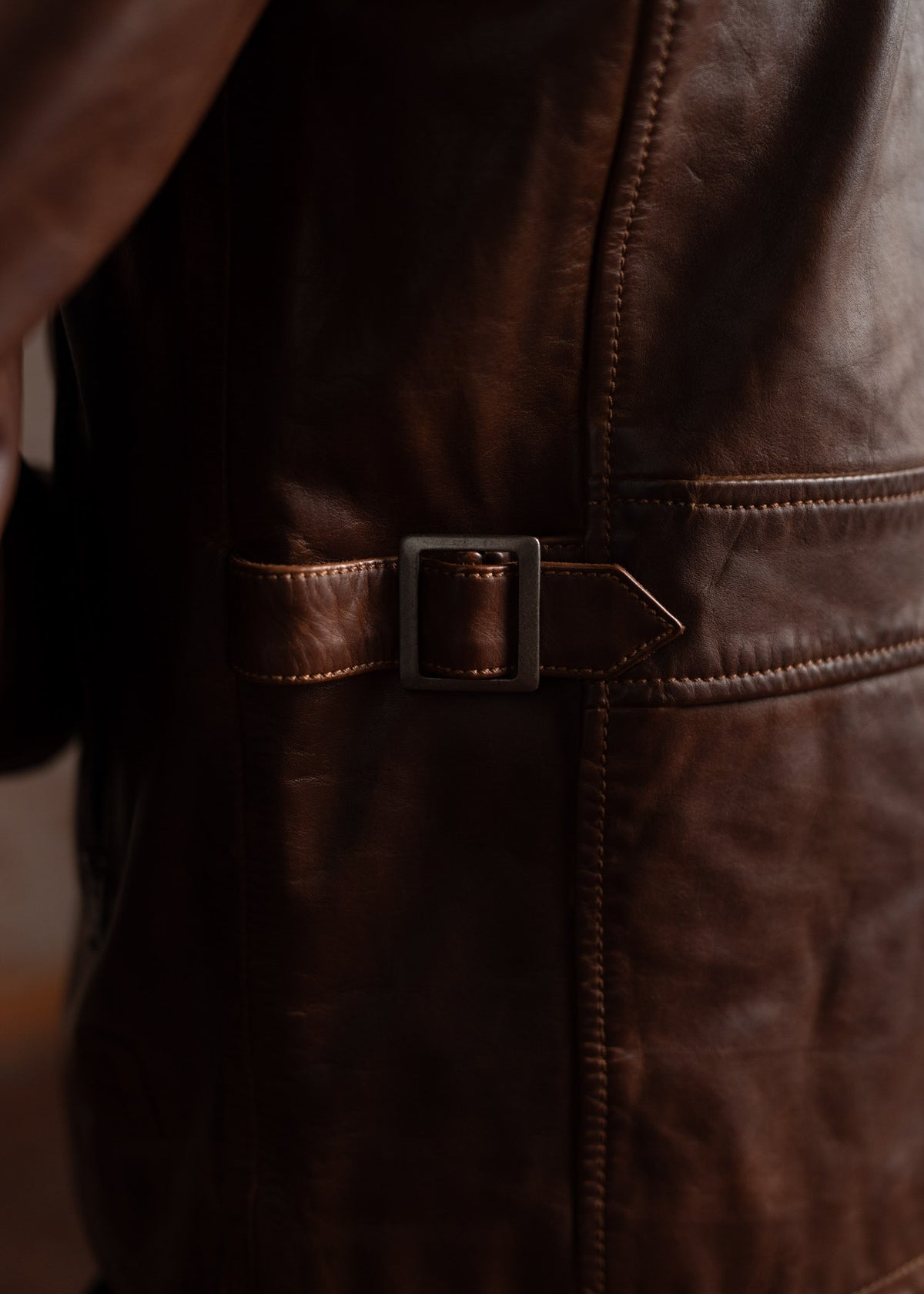 Close-up of a brown leather jacket featuring a metal buckle and detailed stitching, highlighting craftsmanship and texture.