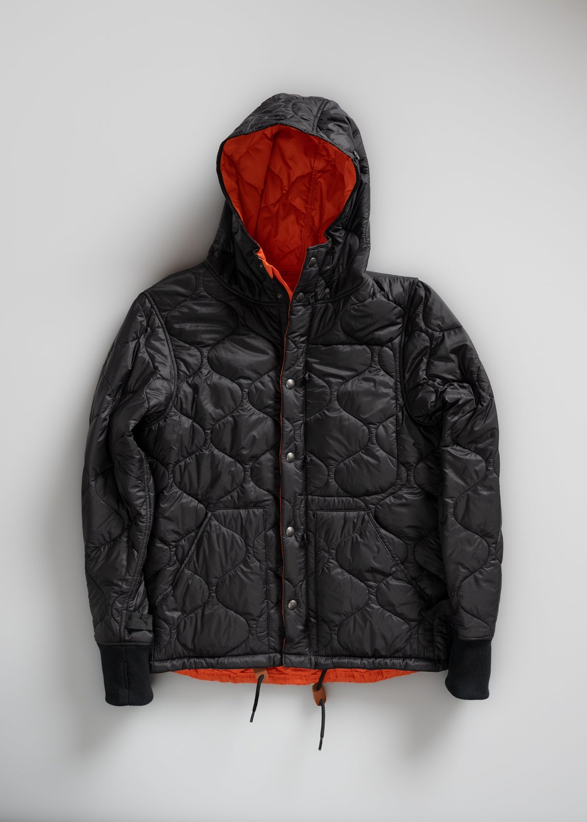 Black quilted jacket with an orange lining and hood, featuring a front button closure and drawstrings, displayed on a white background.