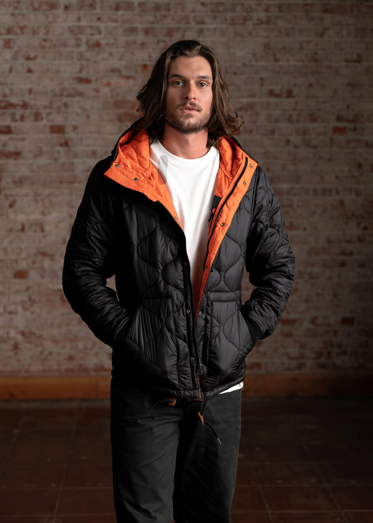 Man in black quilted jacket with orange lining stands against a brick wall, hands in pockets, wearing a white shirt.