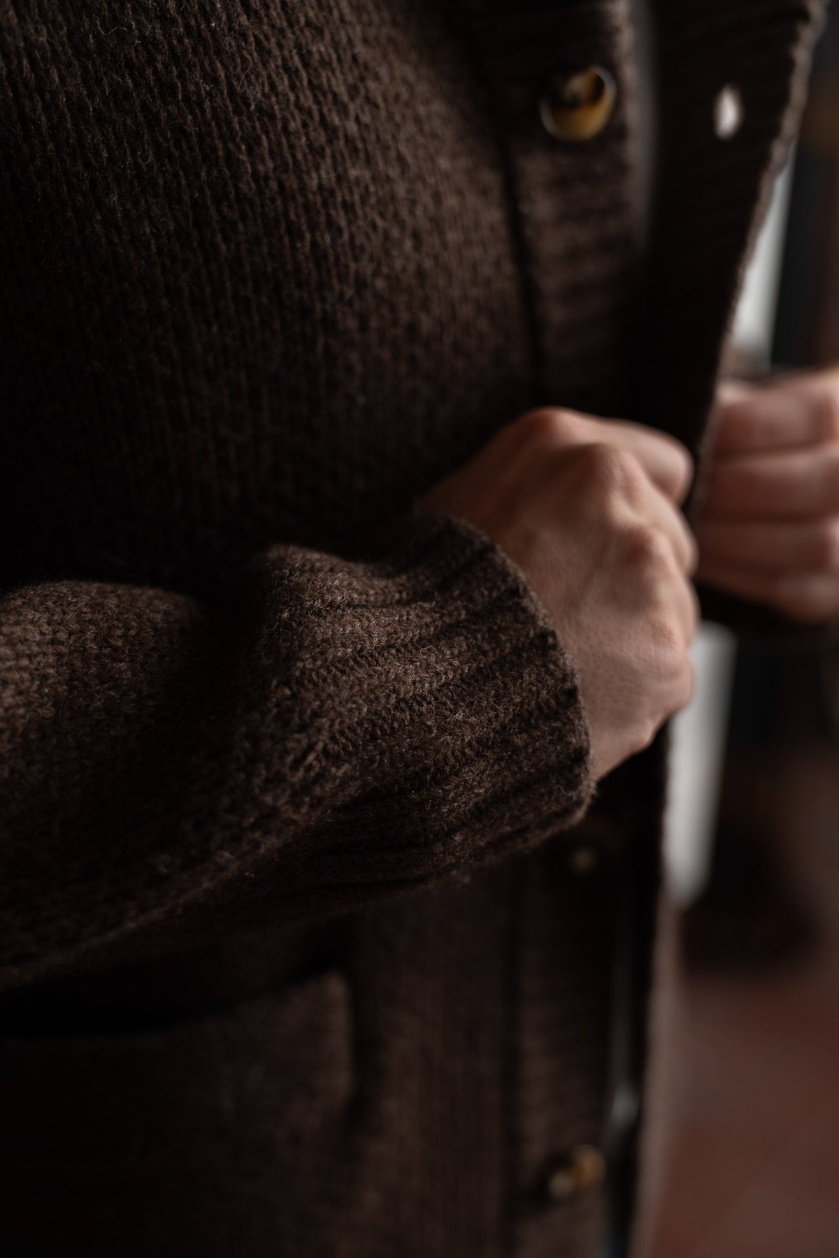 Hands buttoning a cozy, textured brown cardigan, highlighting the knit pattern and warm fabric in soft lighting.