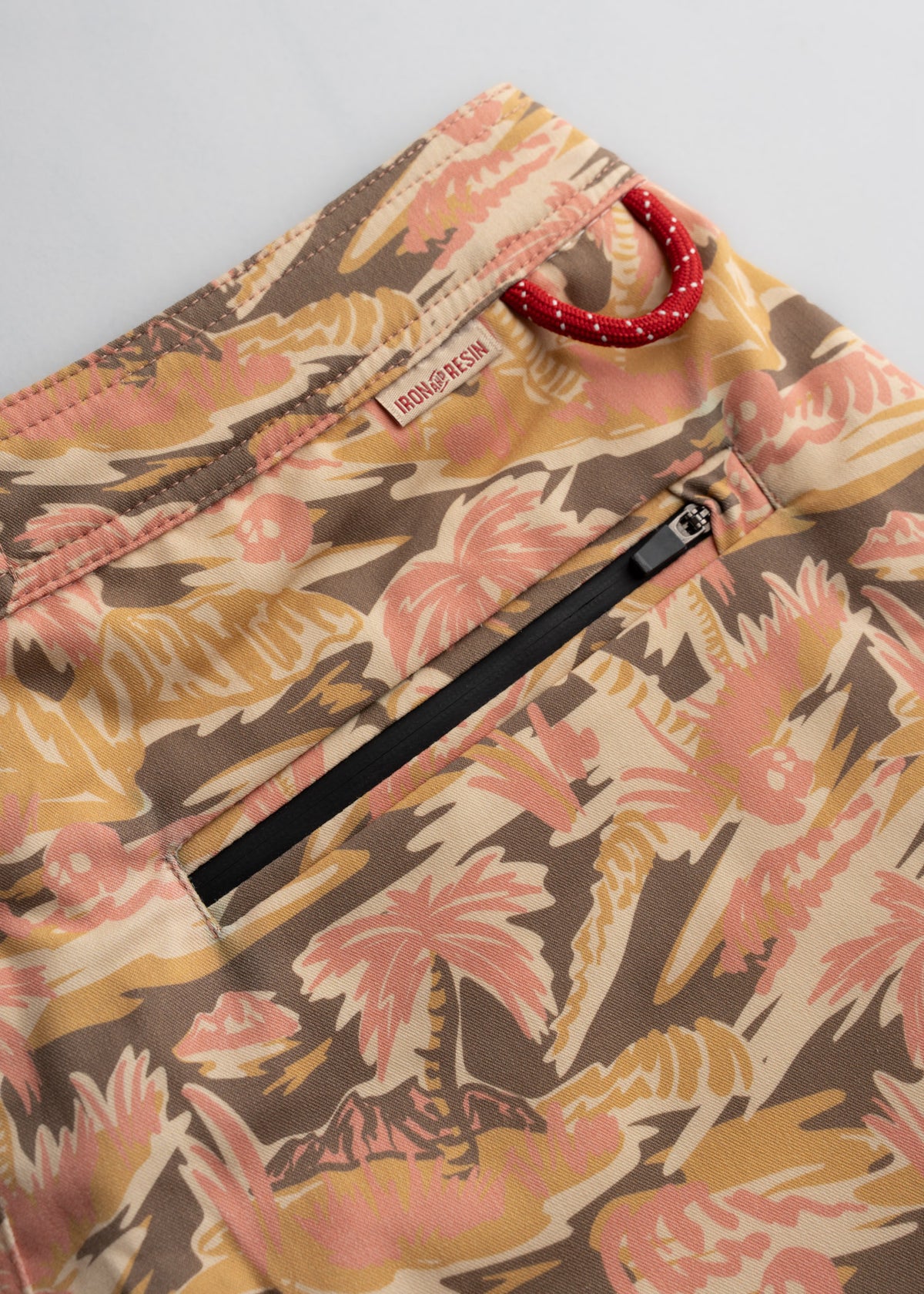 Close-up of tropical-patterned shorts with a zip pocket and red loop. The Iron & Resin label is visible on the waistband.