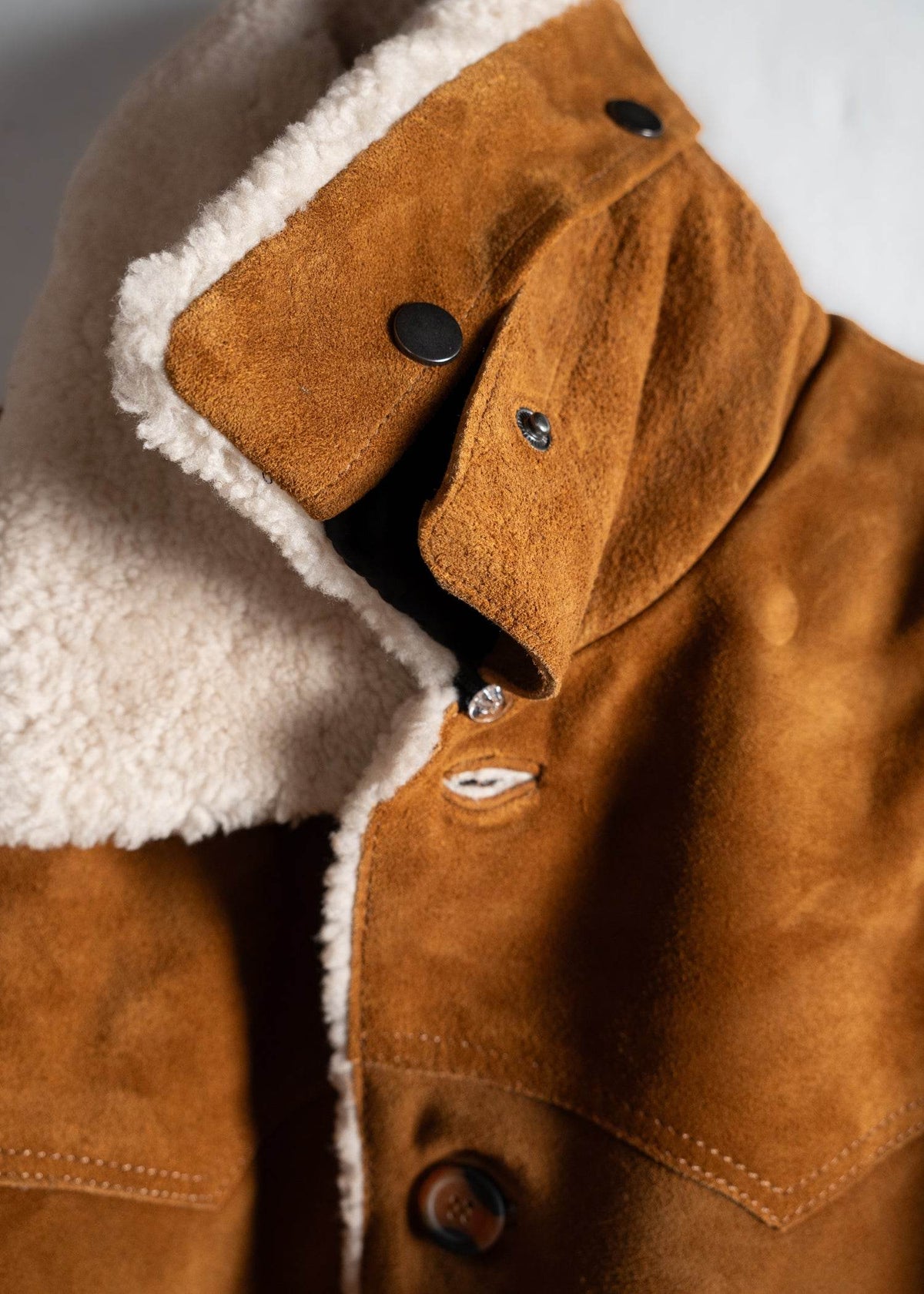 Close-up of a brown suede jacket with a fluffy white shearling collar and black buttons, showcasing its texture and design.