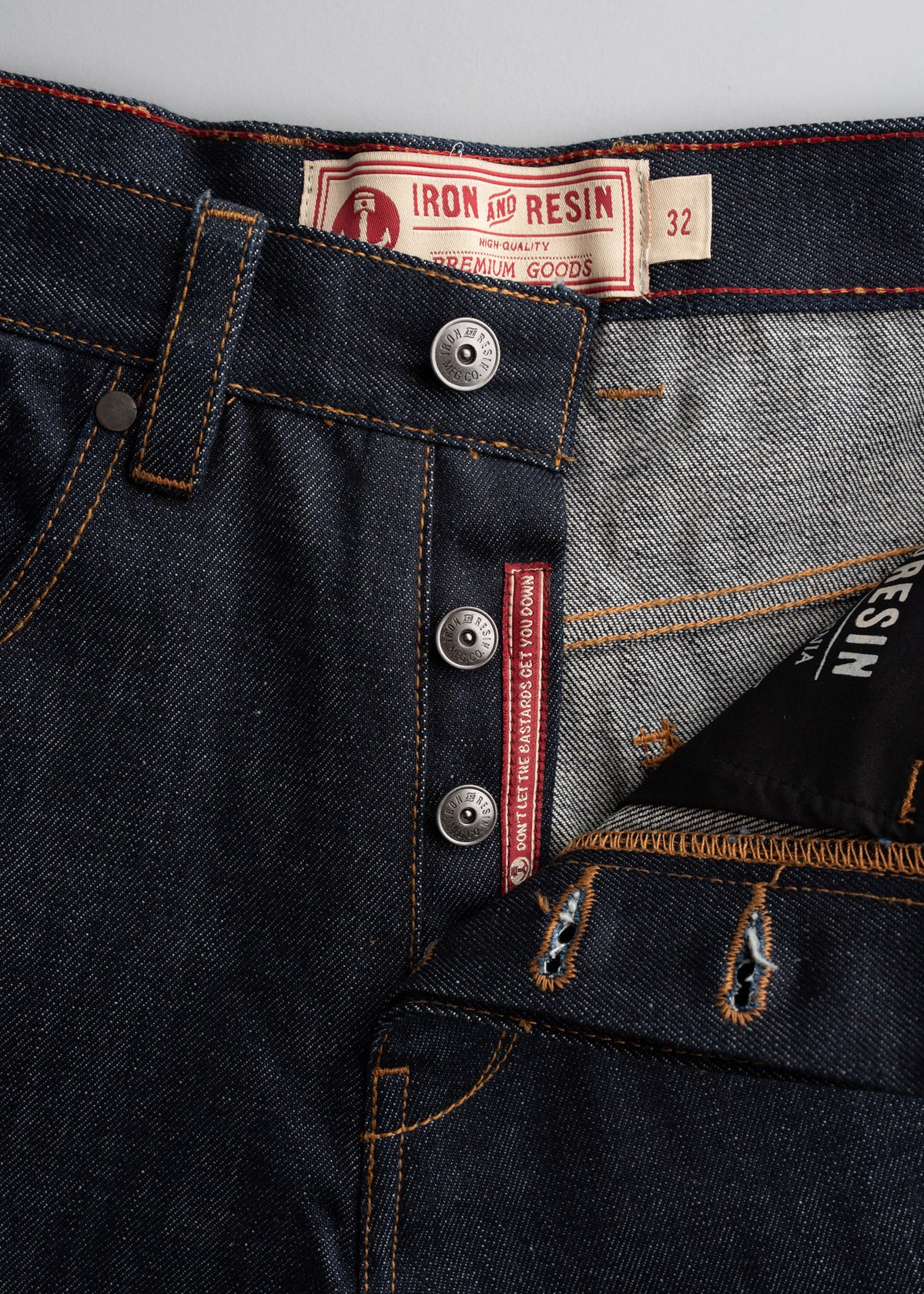 Close-up of Iron and Resin jeans with button fly, detailed stitching, and label showing size 32. Denim texture is visible.