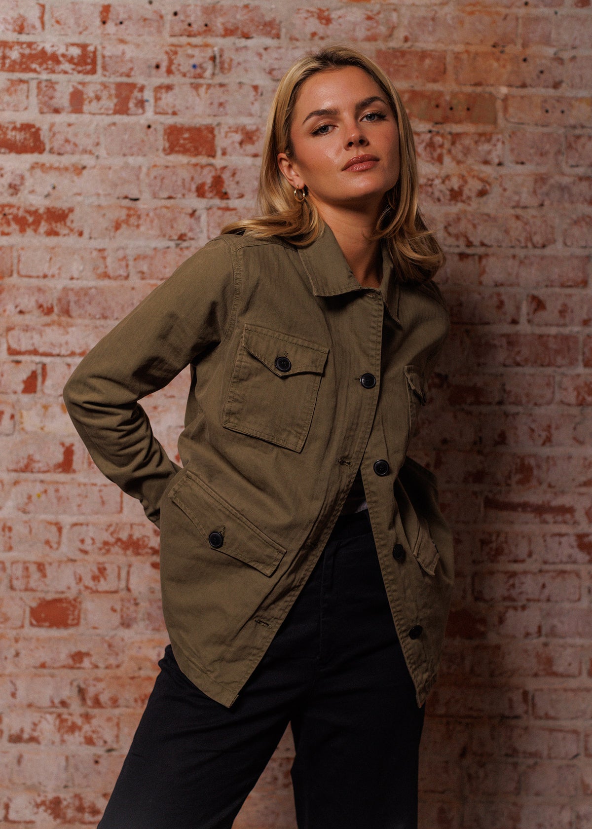 Blonde woman in an olive jacket poses confidently against a rustic brick wall, hands in pockets, exuding casual elegance.