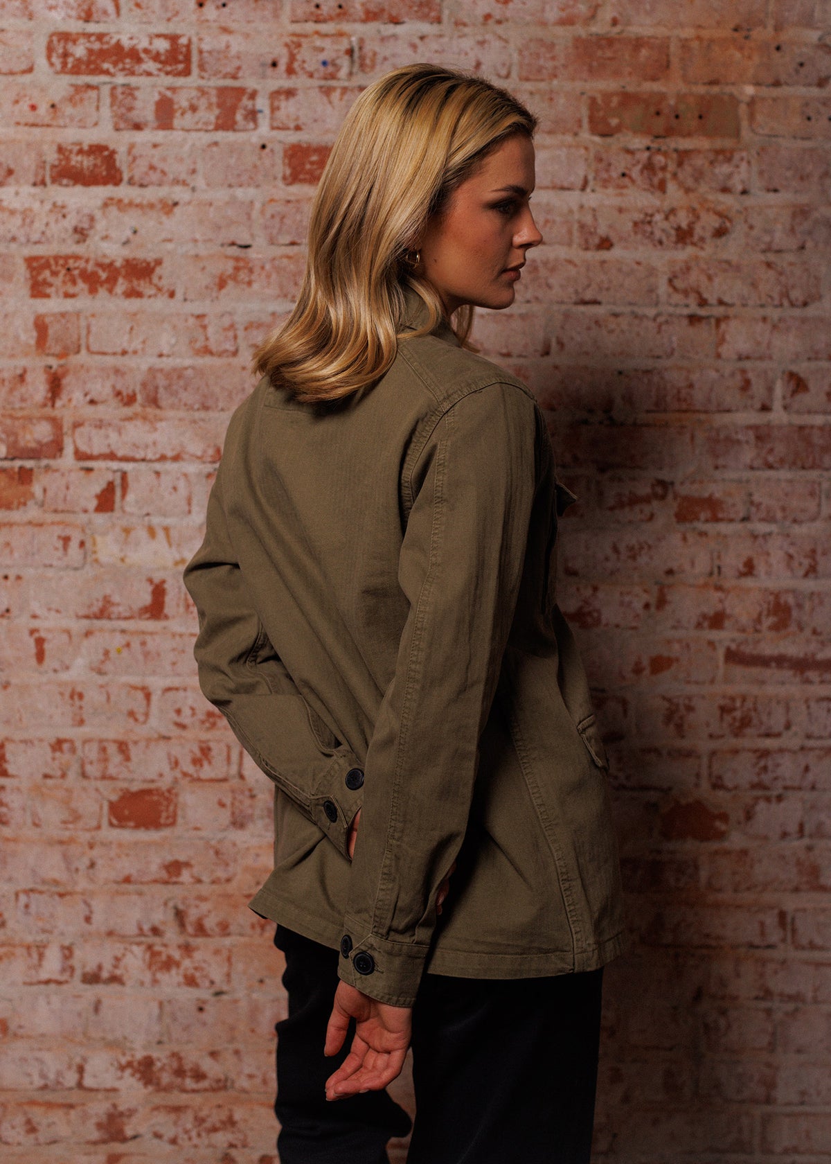 Woman in olive jacket stands against a textured brick wall, looking to the side with hands behind her back, blonde hair flowing.