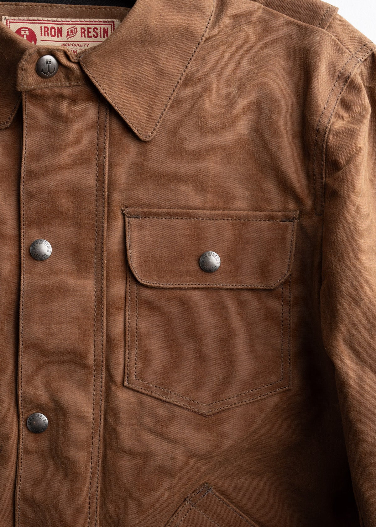 Brown canvas jacket with buttoned flap pocket and collar, featuring Iron and Resin label. Rugged, durable design for outdoor wear.