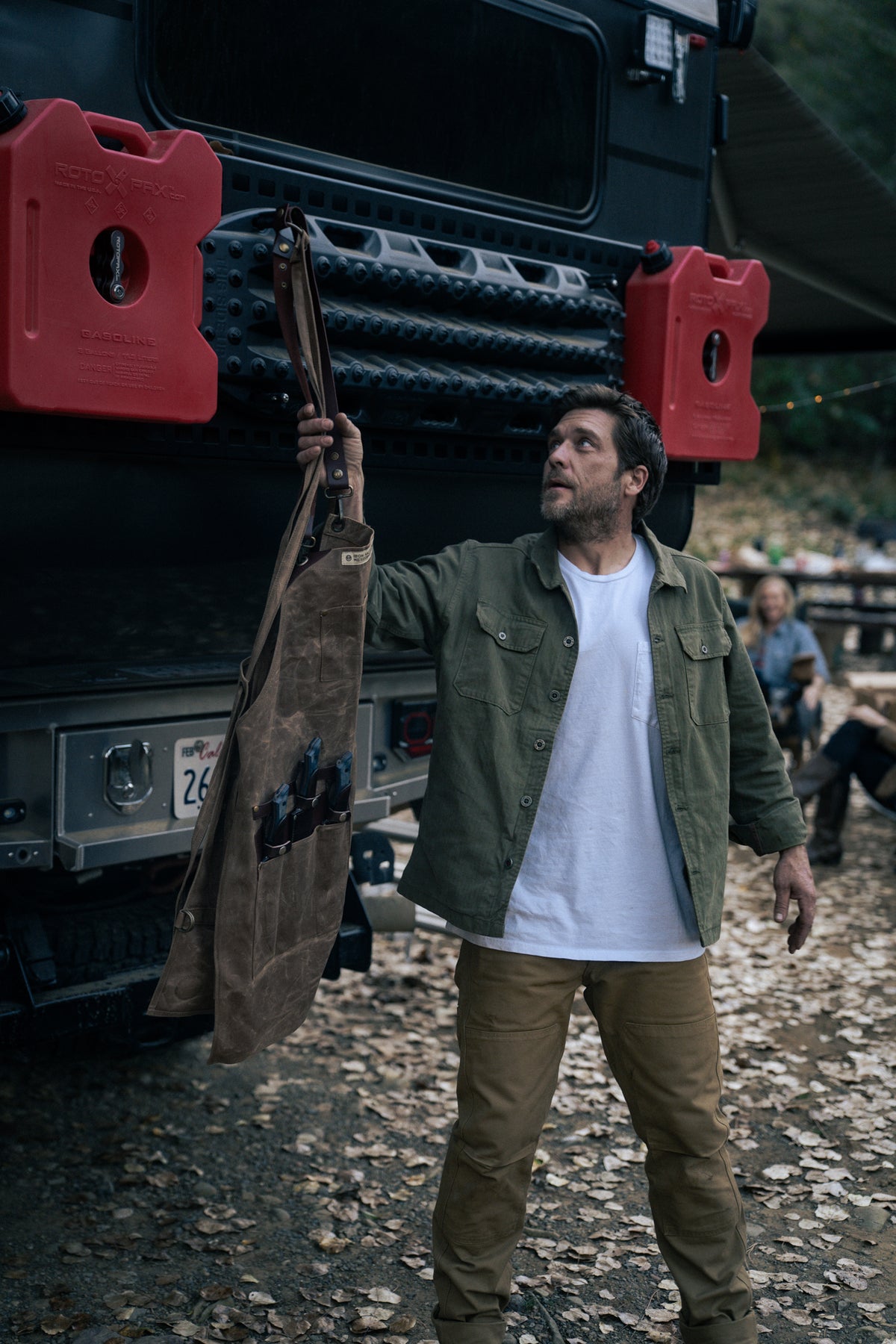 Man in green jacket holds a tool apron by a rugged vehicle with red fuel cans, surrounded by autumn leaves and outdoor setting.