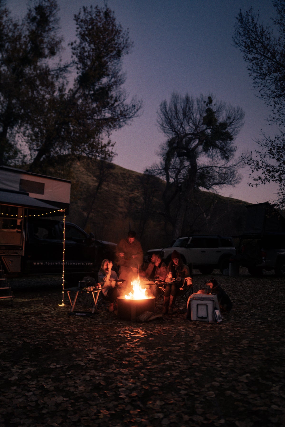 A group of friends sits around a warm campfire at dusk, surrounded by trees and parked vehicles, enjoying an outdoor gathering.