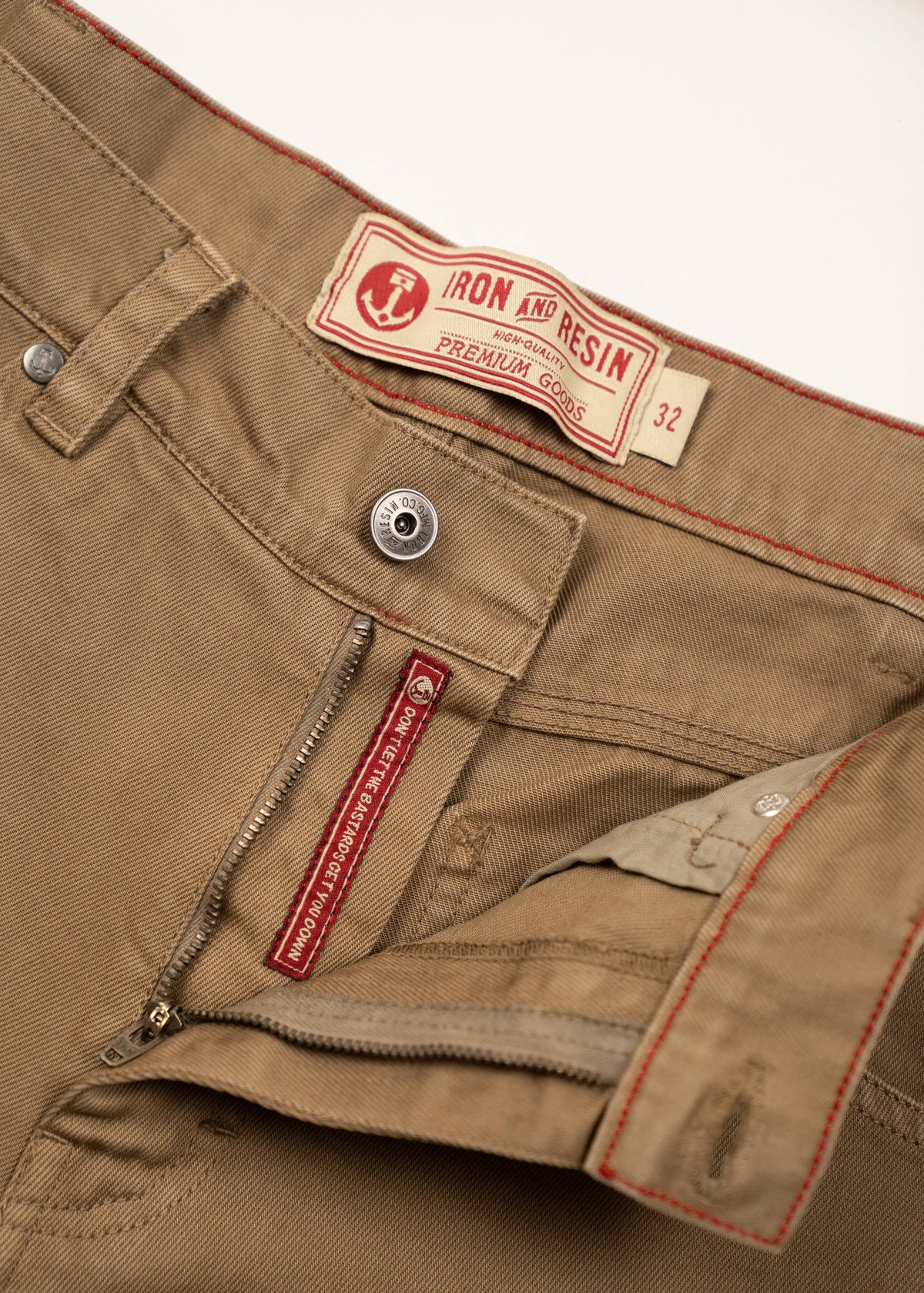 Close-up of khaki pants with Iron and Resin label, size 32. Red stitching accents and zipper detail are visible.