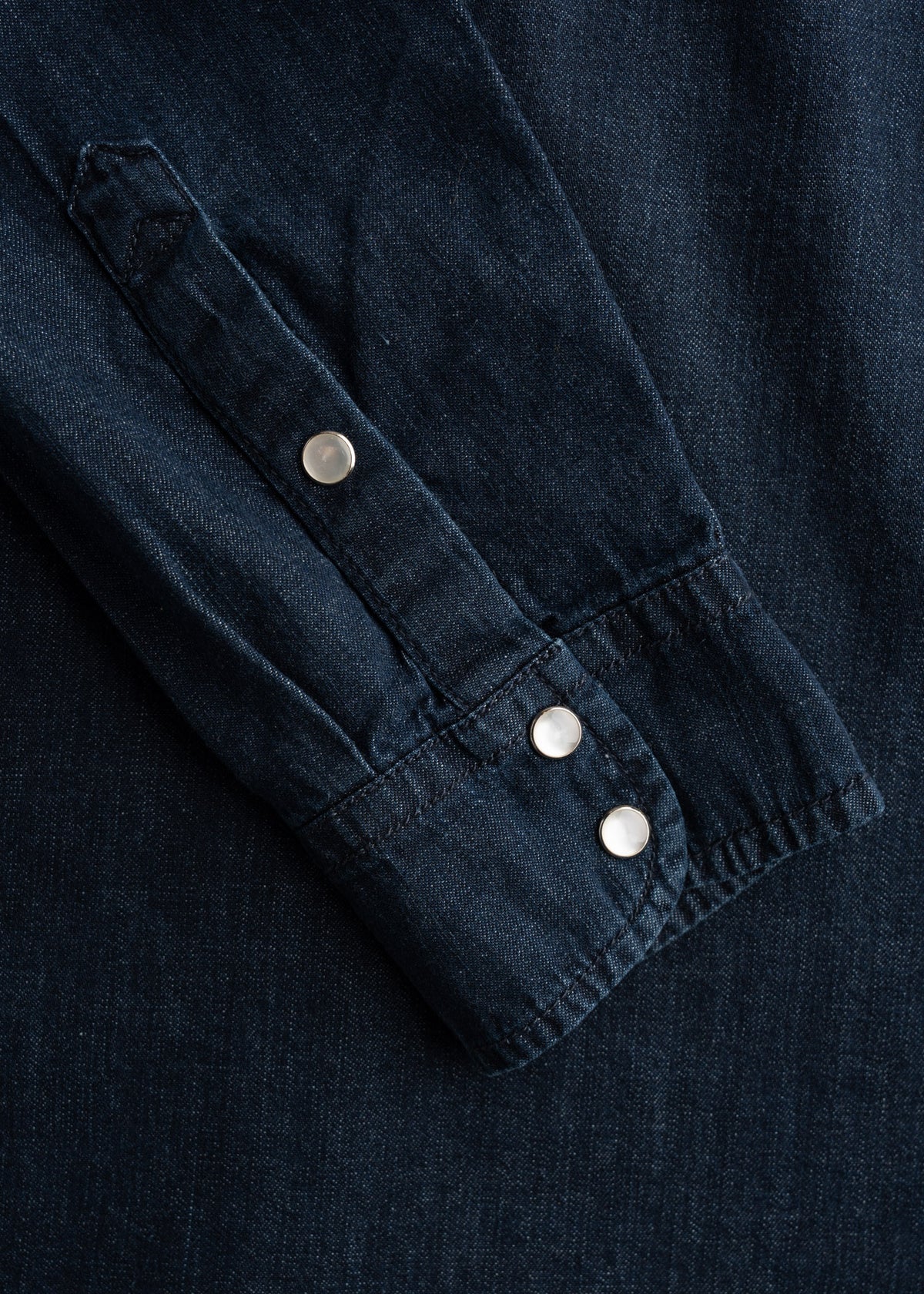 Close-up of a dark denim shirt sleeve with silver snap buttons, showcasing texture and stitching details.