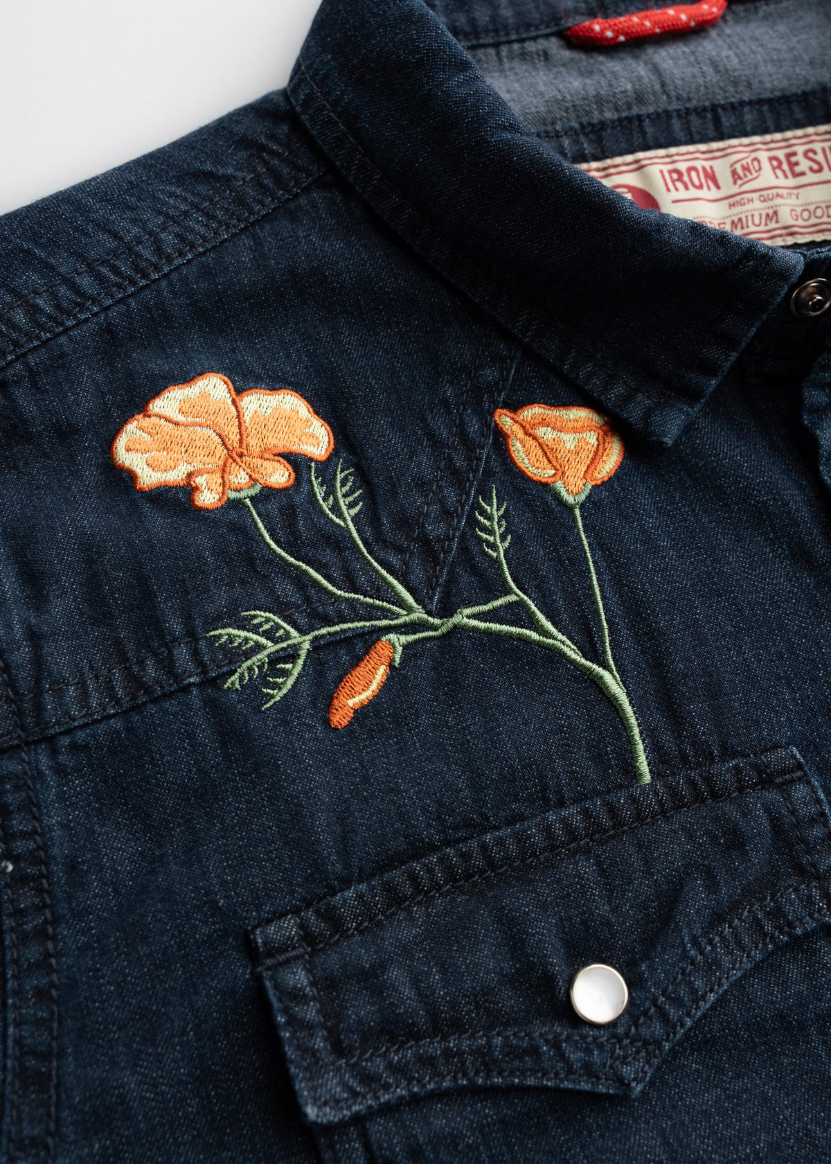 Denim shirt with orange poppy embroidery above pocket, featuring detailed stitching and a visible label inside the collar.
