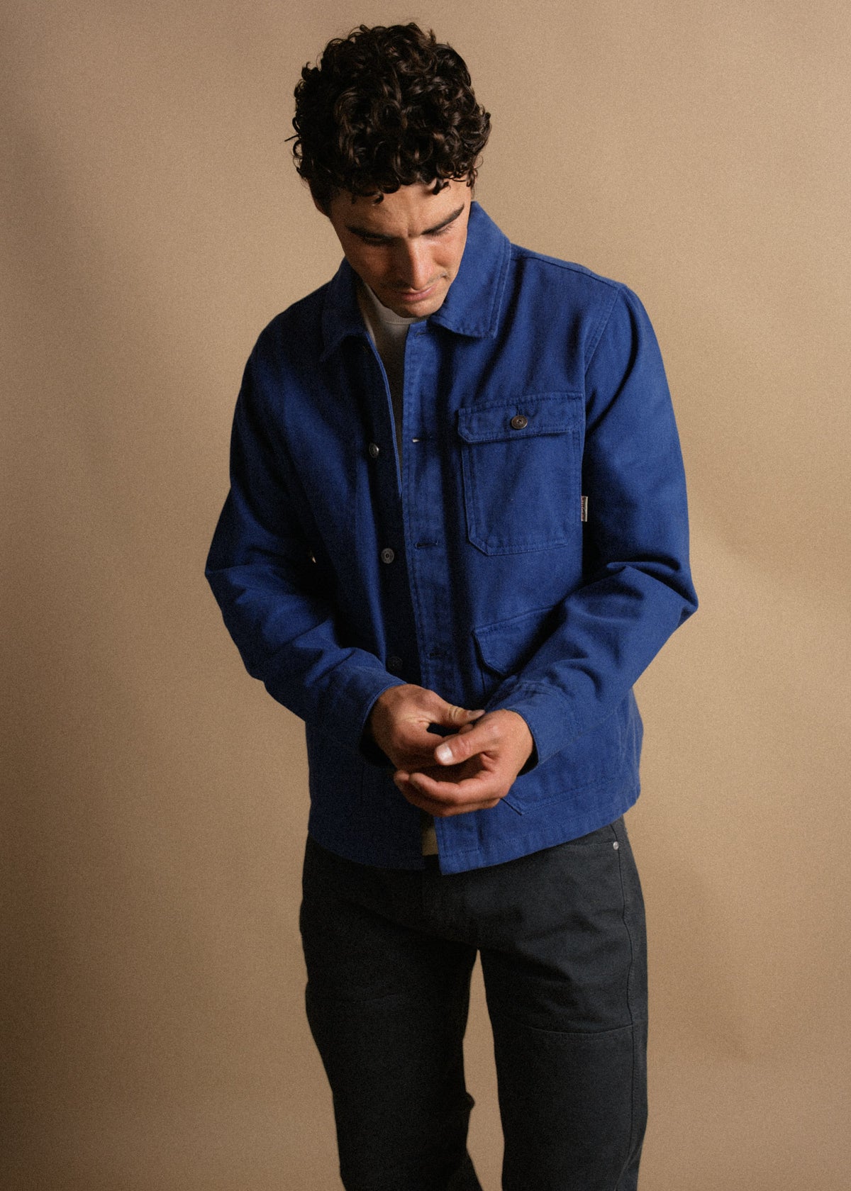 Man in blue jacket looks down, adjusting sleeves. Neutral background highlights casual, stylish outfit.