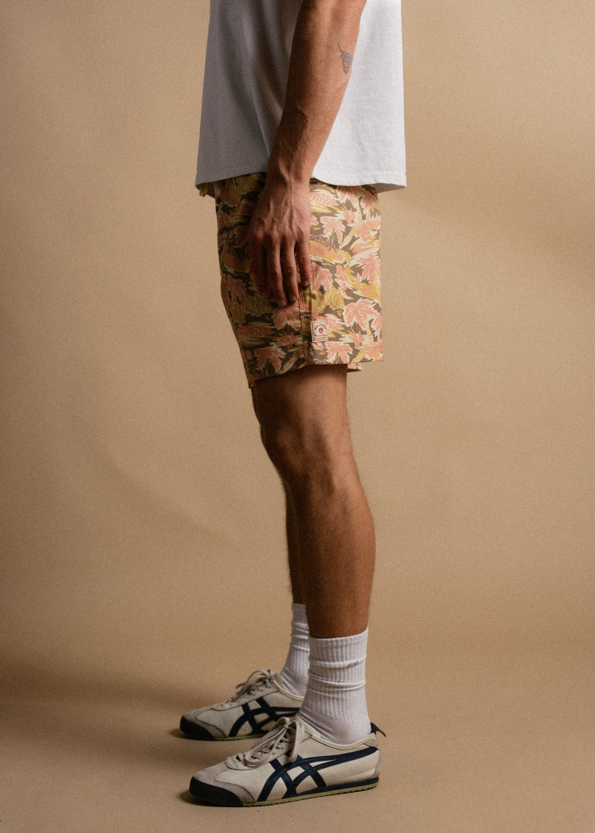 Man in floral shorts and sneakers stands against a beige background, showcasing casual summer fashion with a relaxed vibe.