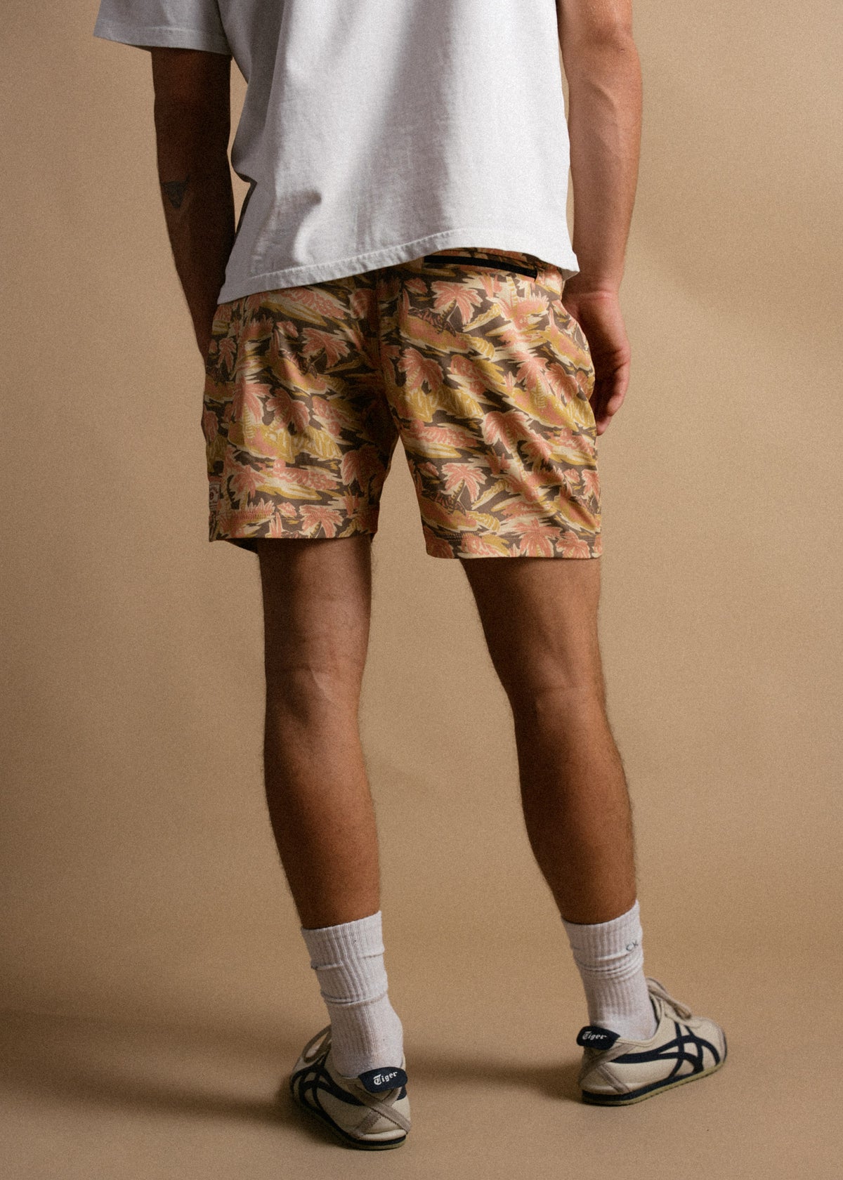 A person in a white shirt and floral shorts stands on a beige background, wearing white socks and sneakers, facing away.