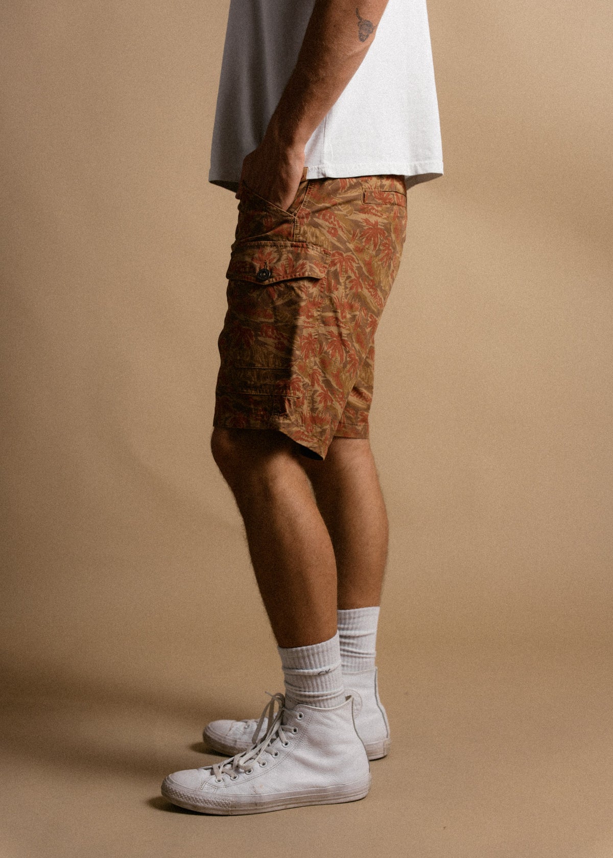 Man in white sneakers, socks, and floral cargo shorts, standing sideways against a beige background, wearing a white t-shirt.