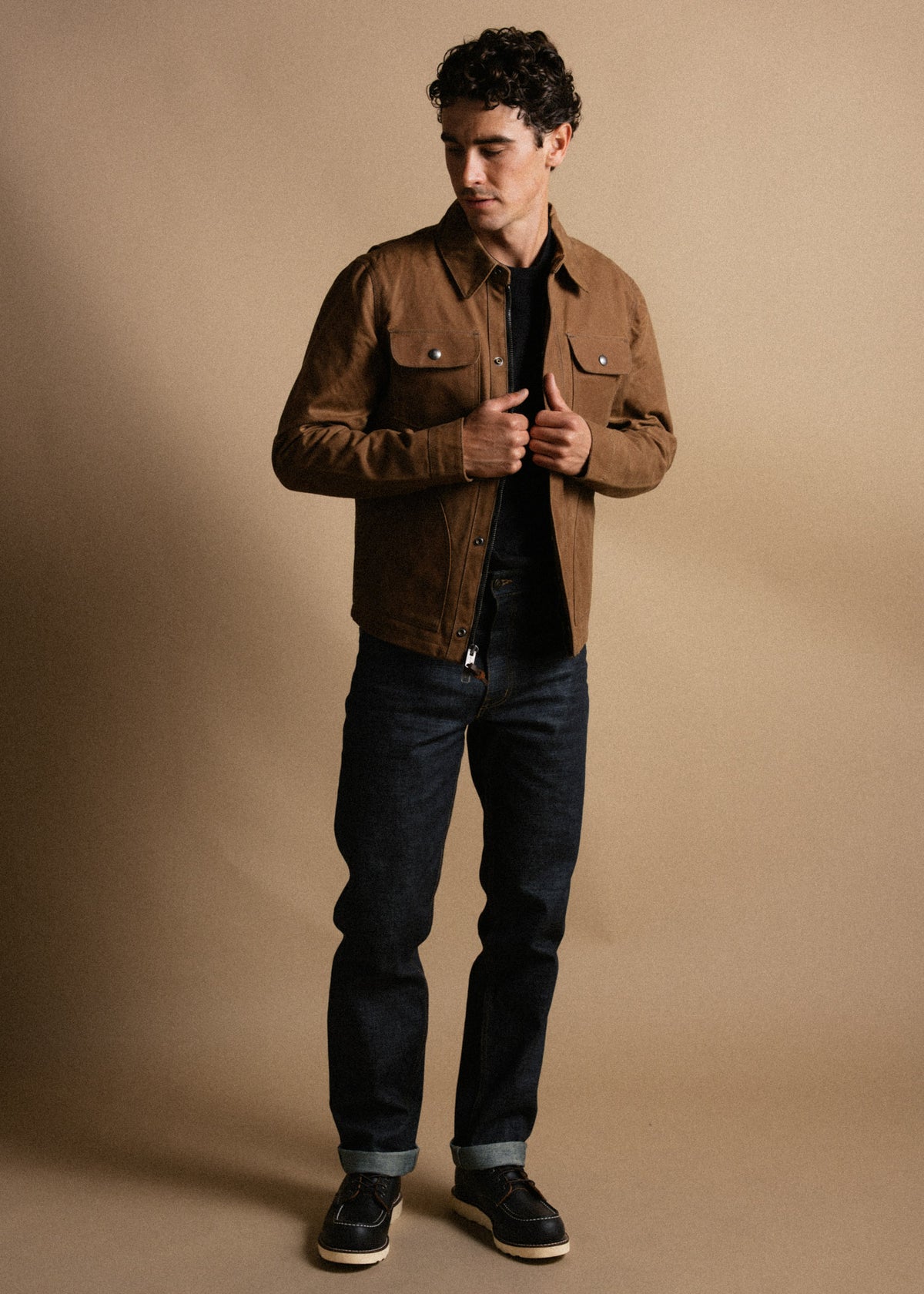 Man in brown jacket and dark jeans stands against a beige background, looking down with hands adjusting his jacket.