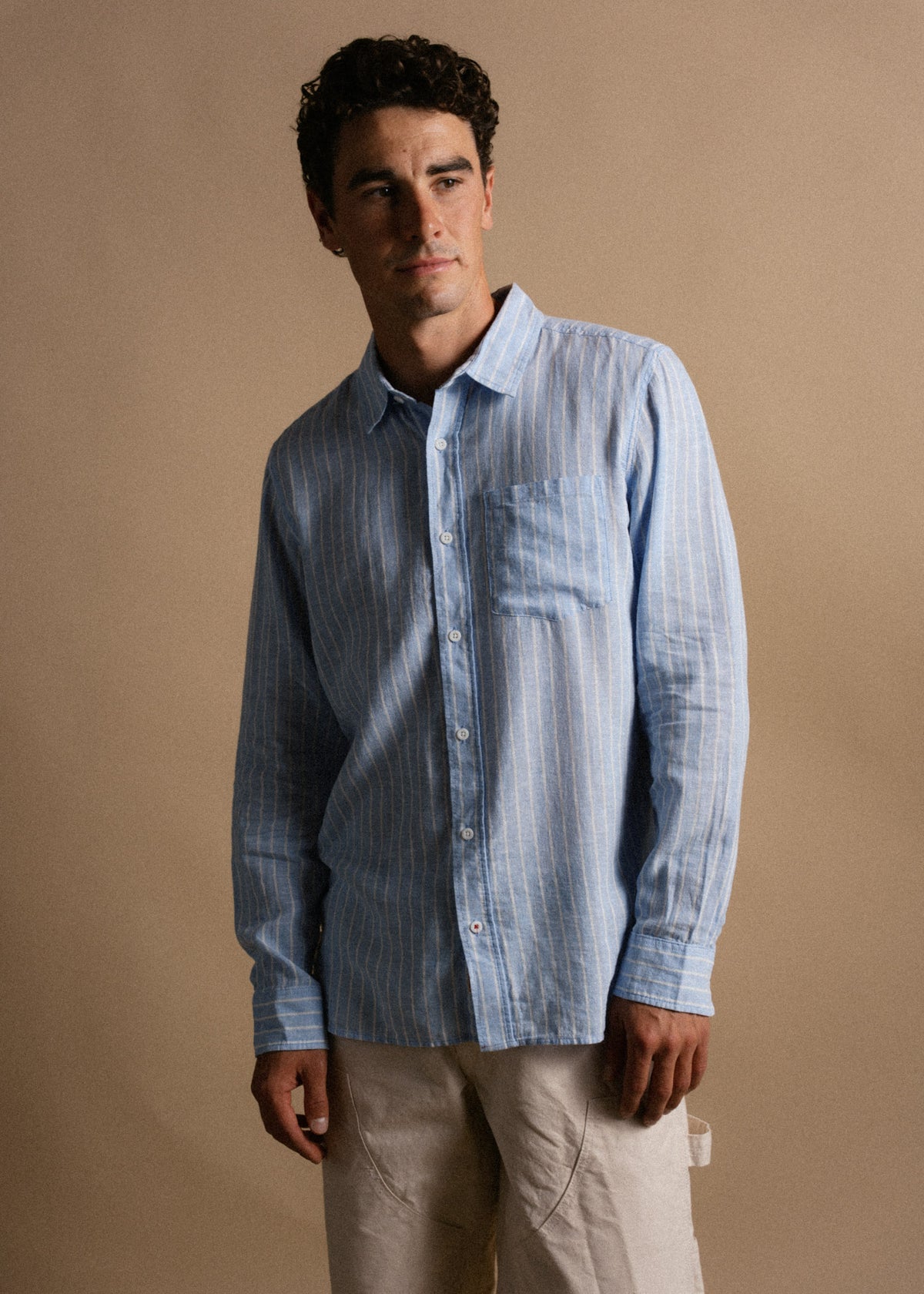 Man in a blue striped shirt and beige pants stands against a neutral background, looking to the side with a thoughtful expression.