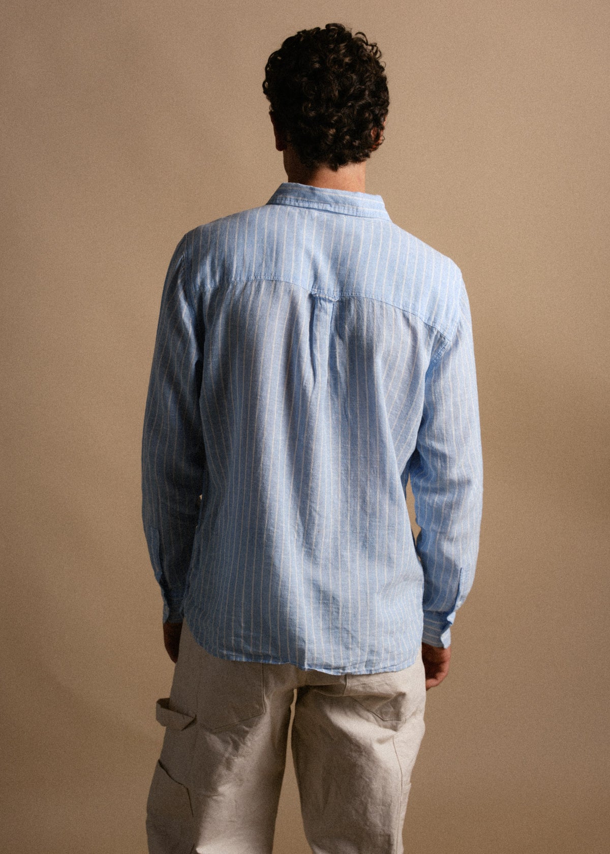 Man in a blue striped shirt and beige pants stands with his back to the camera against a plain background.