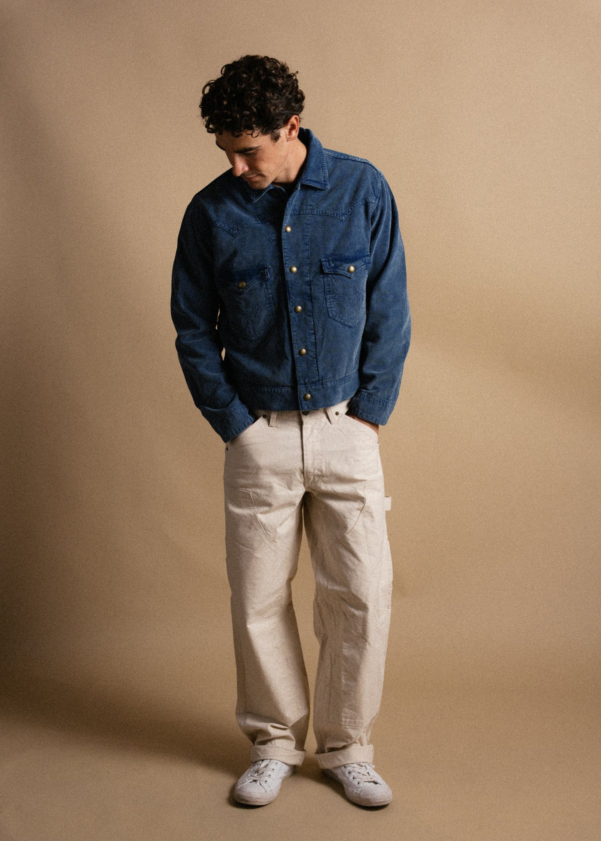 Man in blue denim jacket and beige pants looks down, standing against a plain tan background, hands in pockets, casual style.