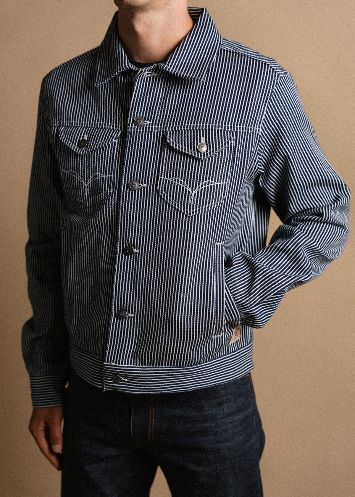 Man in a blue striped denim jacket with buttoned pockets stands against a beige background, showcasing casual fashion style.
