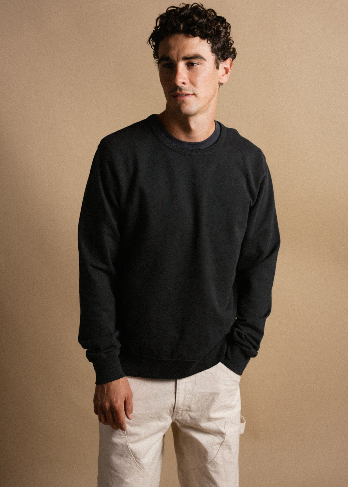 Man in black sweatshirt and white pants stands against a beige background, looking to the side with a neutral expression.
