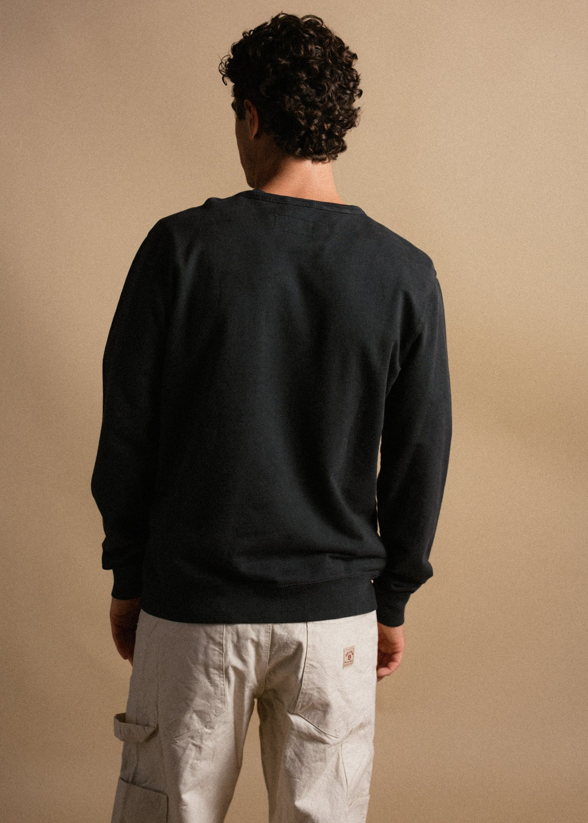 Man in black sweater and beige cargo pants stands with back to camera against a plain background, showcasing casual style.