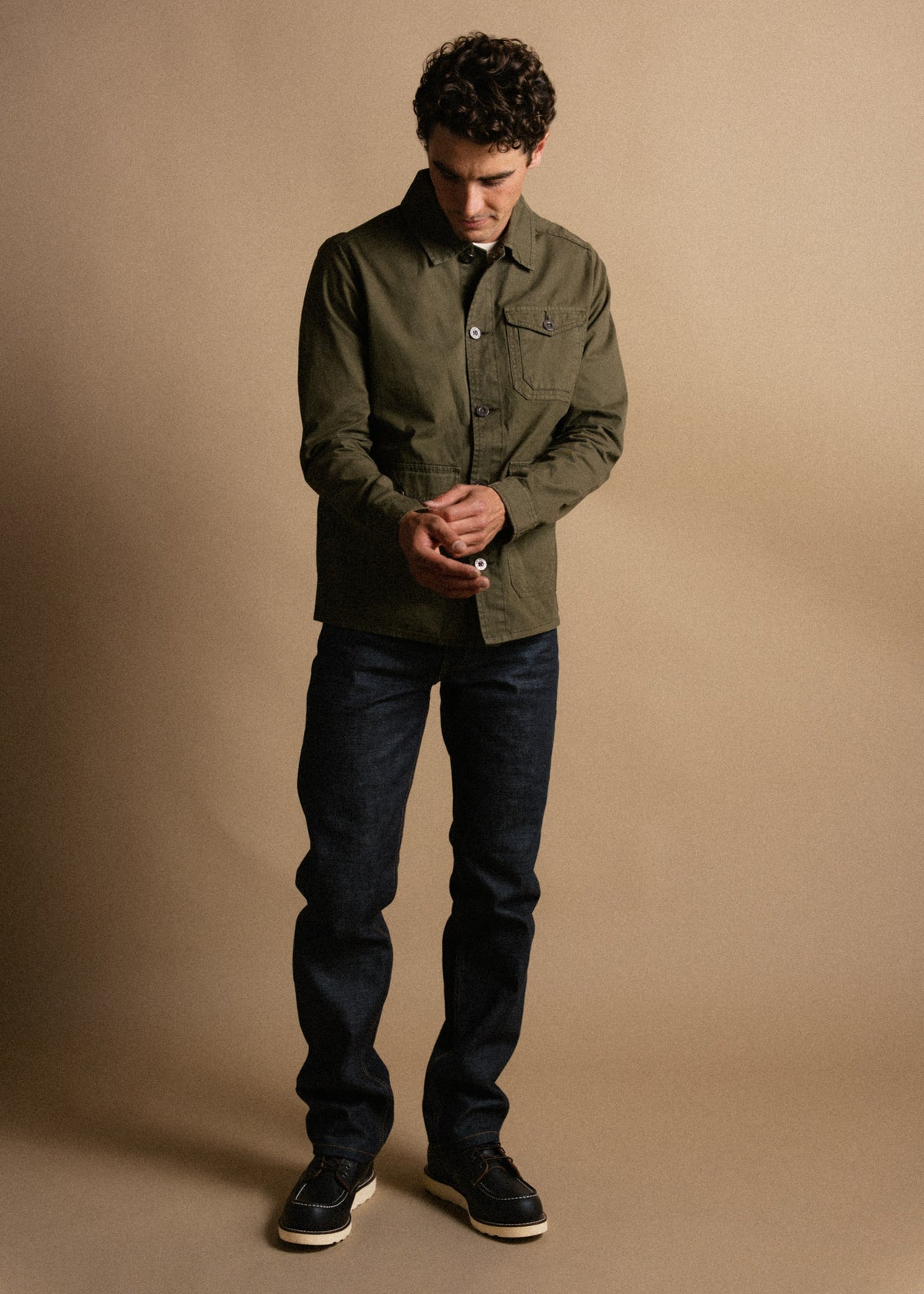 Man in olive jacket and dark jeans stands against beige background, adjusting cuffs. Casual, stylish look with focus on outfit.