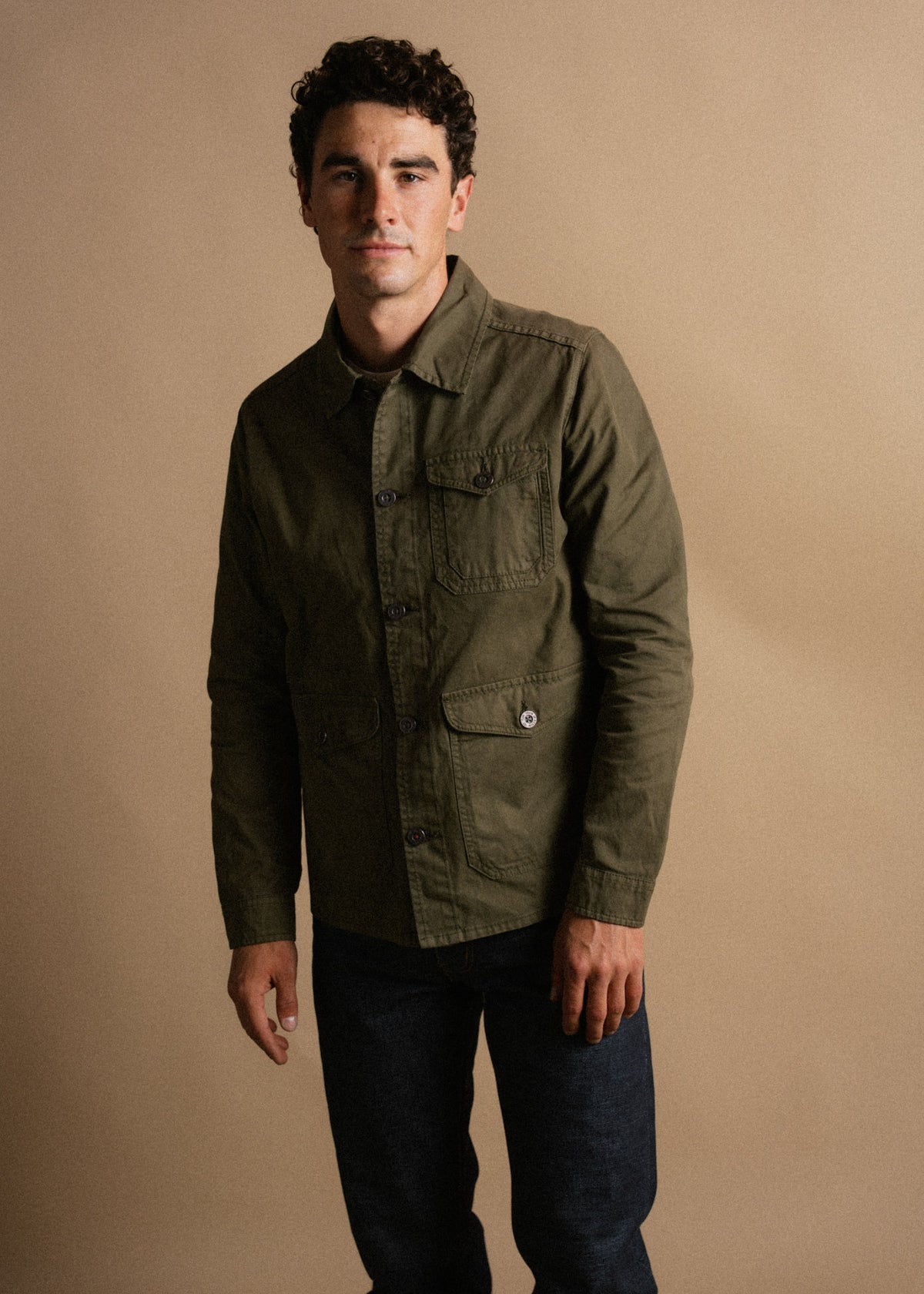 Man in olive jacket and jeans stands against a beige background, exuding a casual yet stylish look with a confident expression.