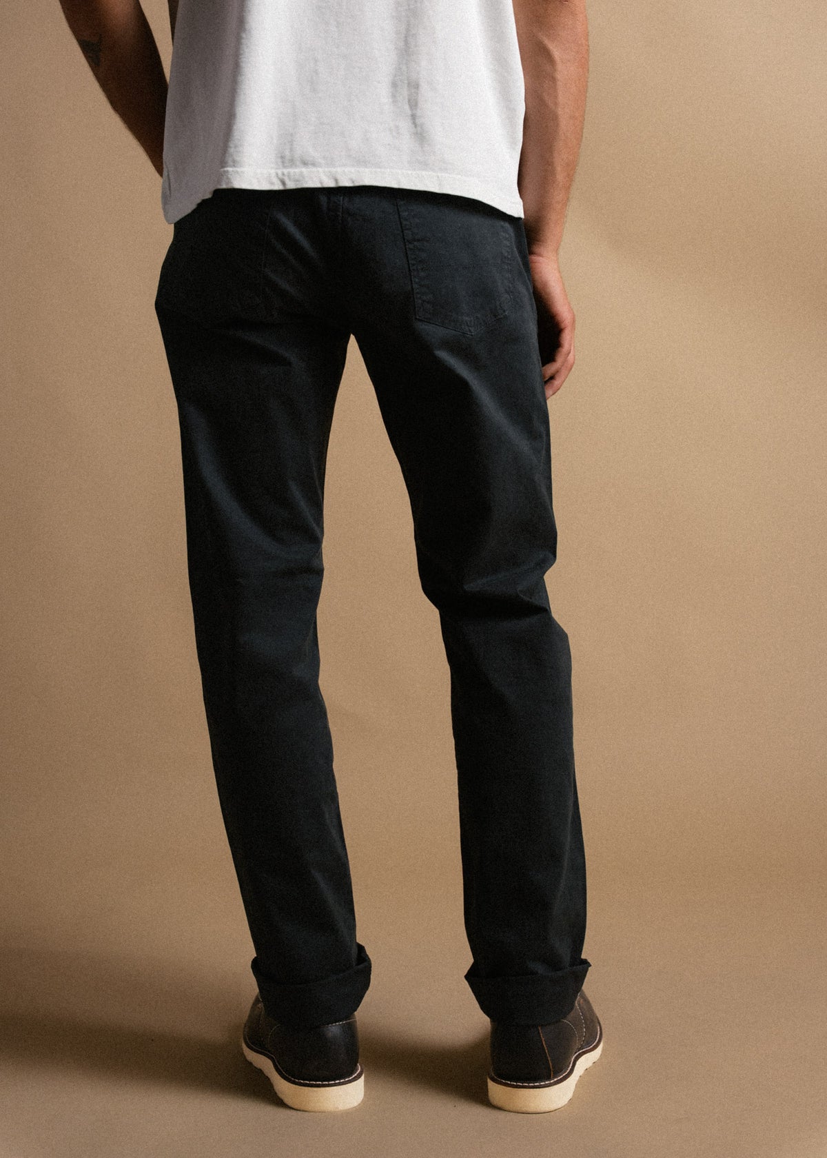 Man in black pants and white shirt stands on a beige background, showcasing casual style with rolled cuffs and brown shoes.