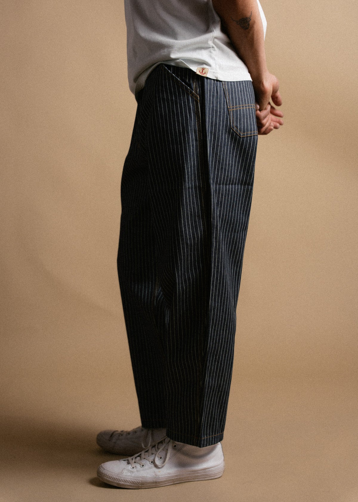 Person in pinstripe pants and white sneakers stands against a beige background, hands clasped behind back, wearing a white shirt.