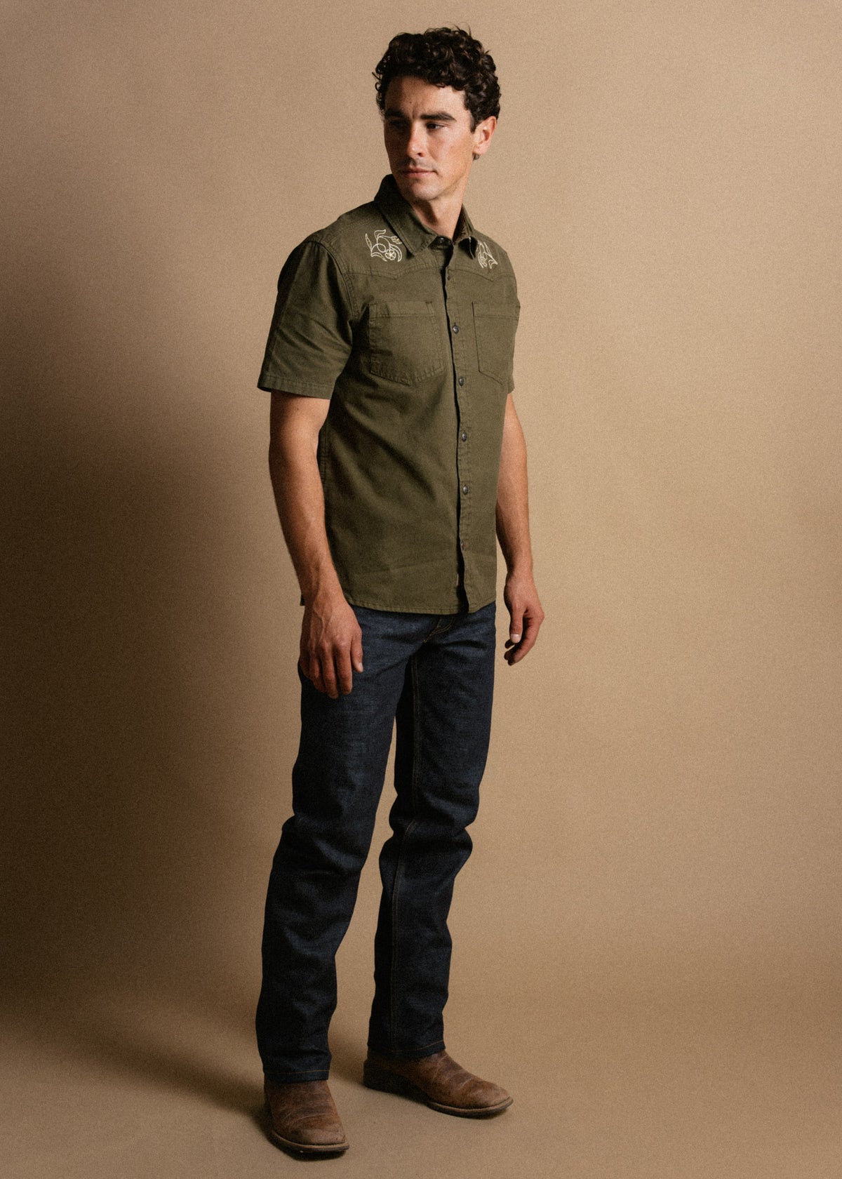 Man in olive shirt and jeans stands against a beige background, looking to the side, embodying casual style and confidence.