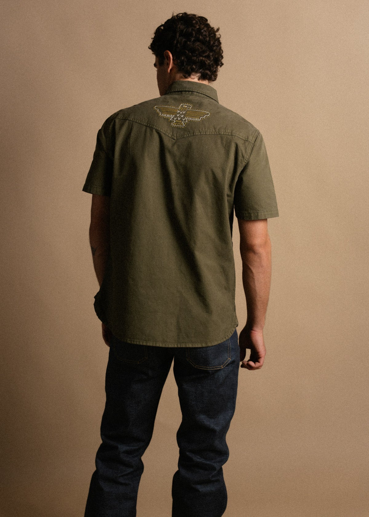 Man in olive shirt with eagle embroidery stands against a beige background, facing away, wearing dark jeans.