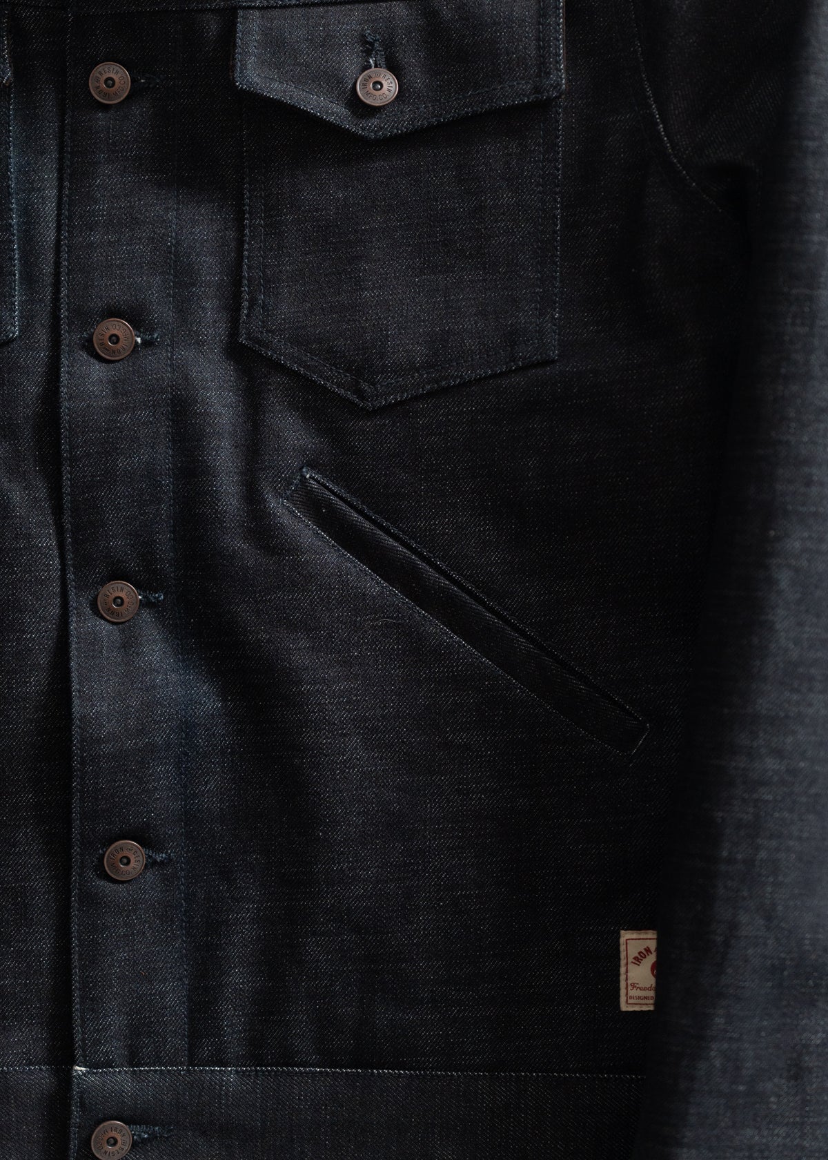 Dark denim jacket with brown buttons, chest pockets, and a visible label. The fabric texture is prominently displayed.