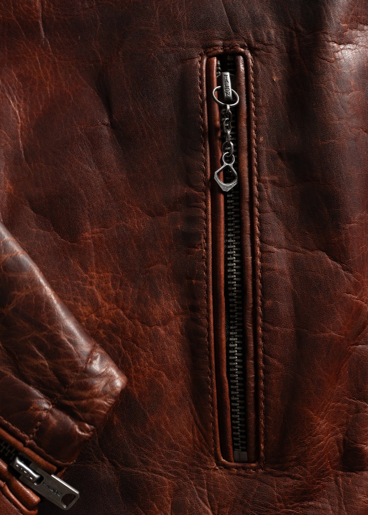 Close-up of a brown leather jacket's textured surface, highlighting a vertical zipper pocket with a metallic pull tab.