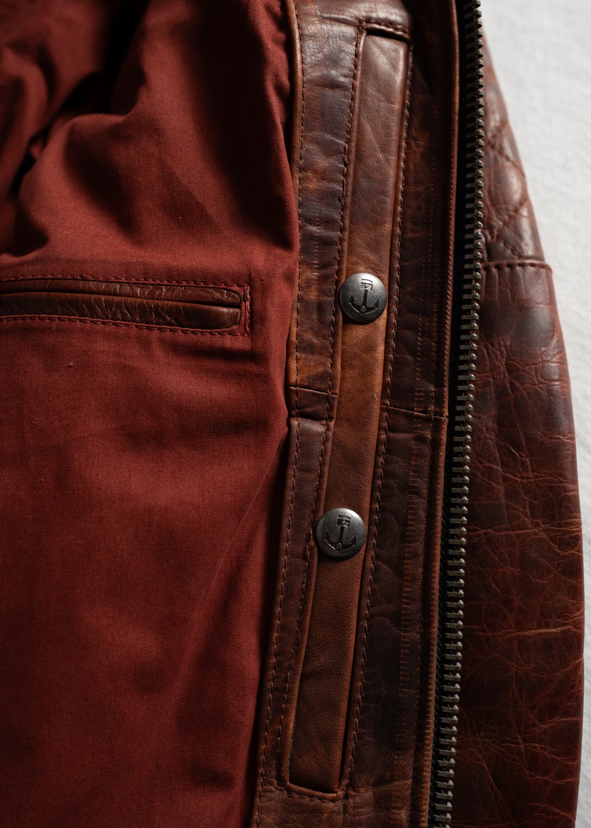 Close-up of a brown leather jacket's interior, showing red lining, a pocket, and anchor-embossed buttons near the zipper.