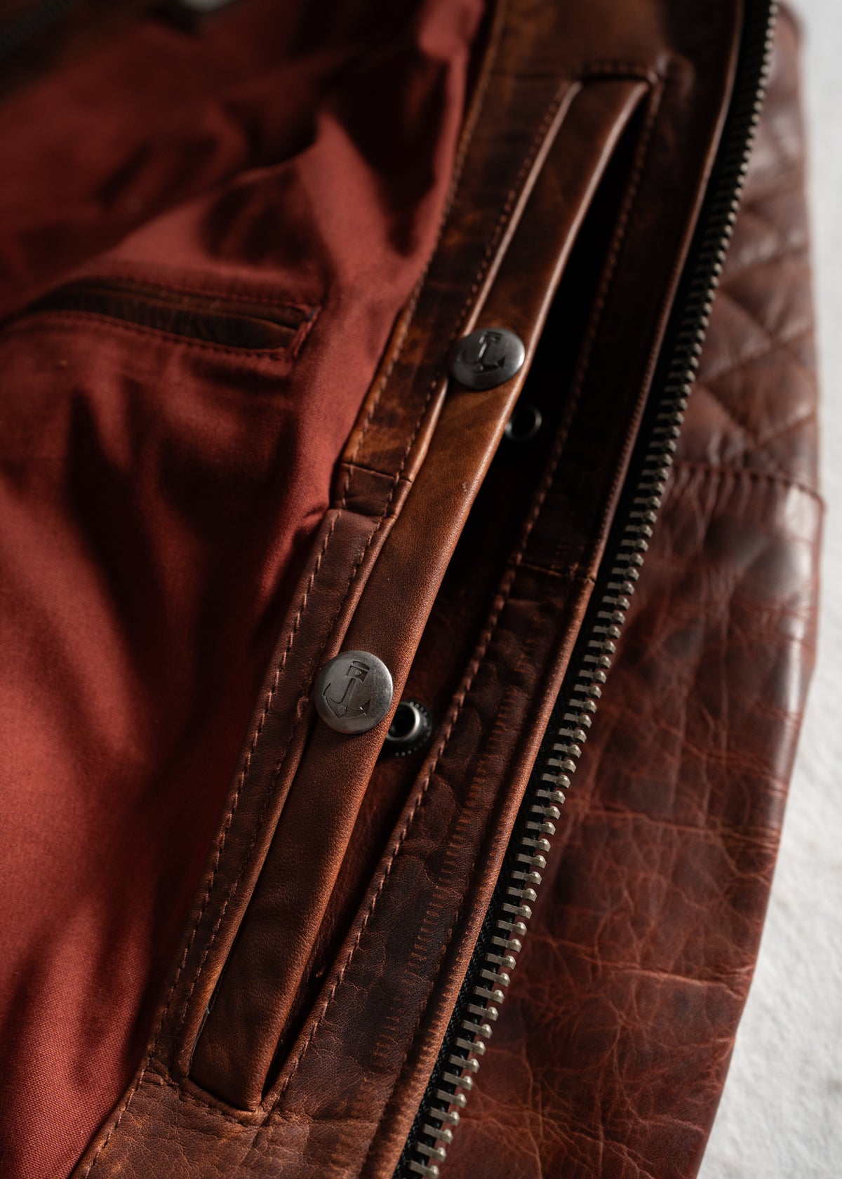 Close-up of a brown leather jacket with a visible zipper and button detail, showcasing its rich texture and red fabric lining.