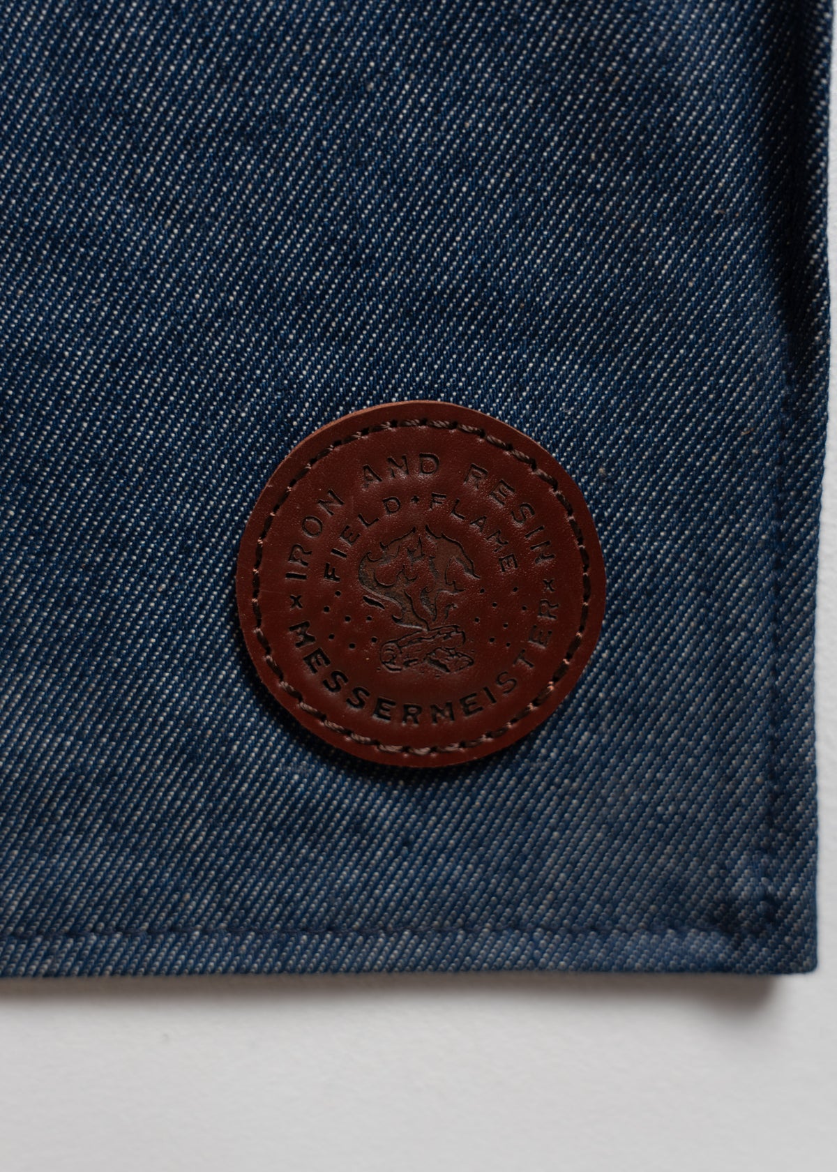 Denim fabric with a brown leather patch embossed with "Iron and Resin" and "Messermeister" logos, featuring a flame design.
