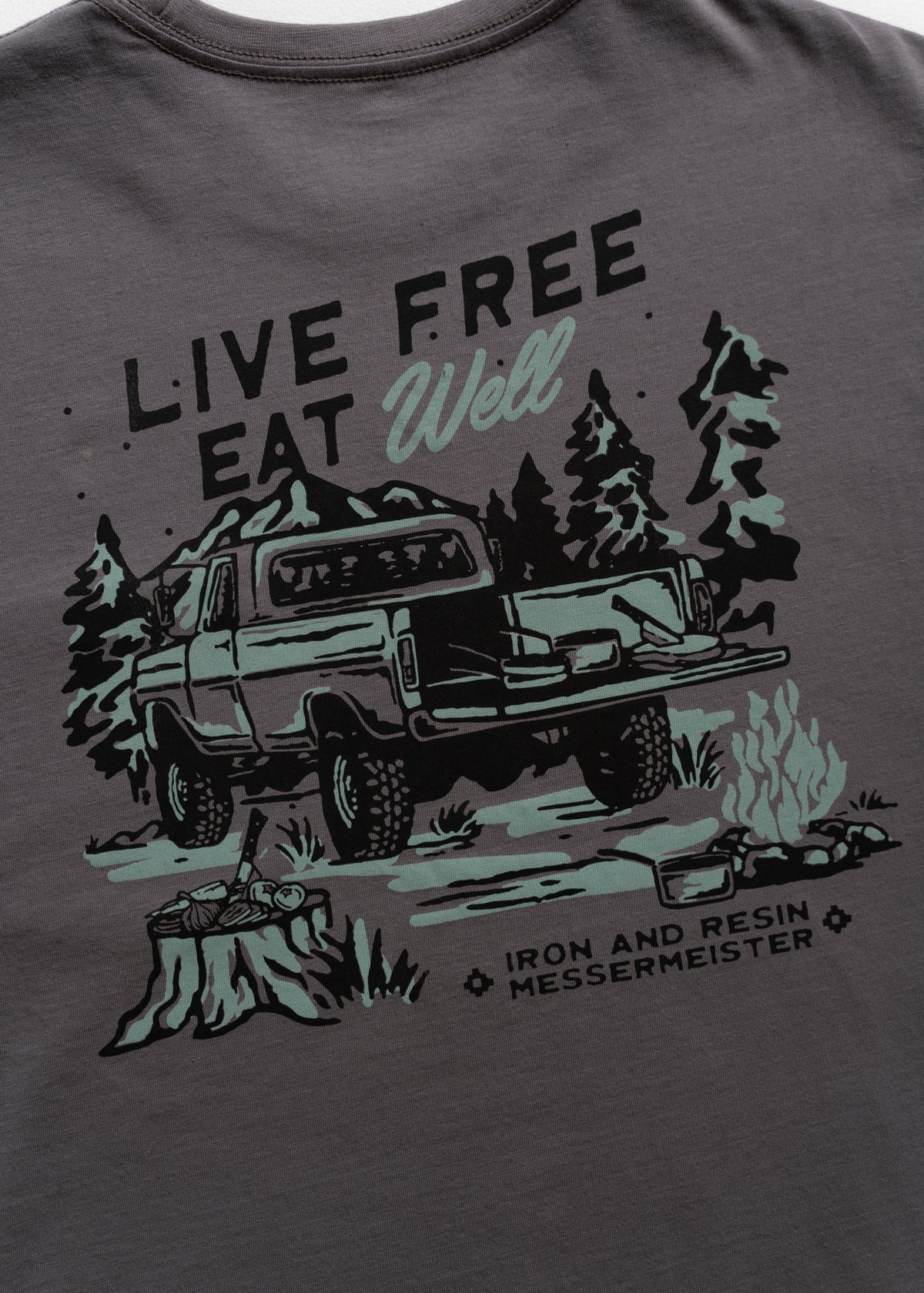 T-shirt design with a truck, trees, and campfire, featuring "Live Free Eat Well" and "Iron and Resin Messermeister" text.