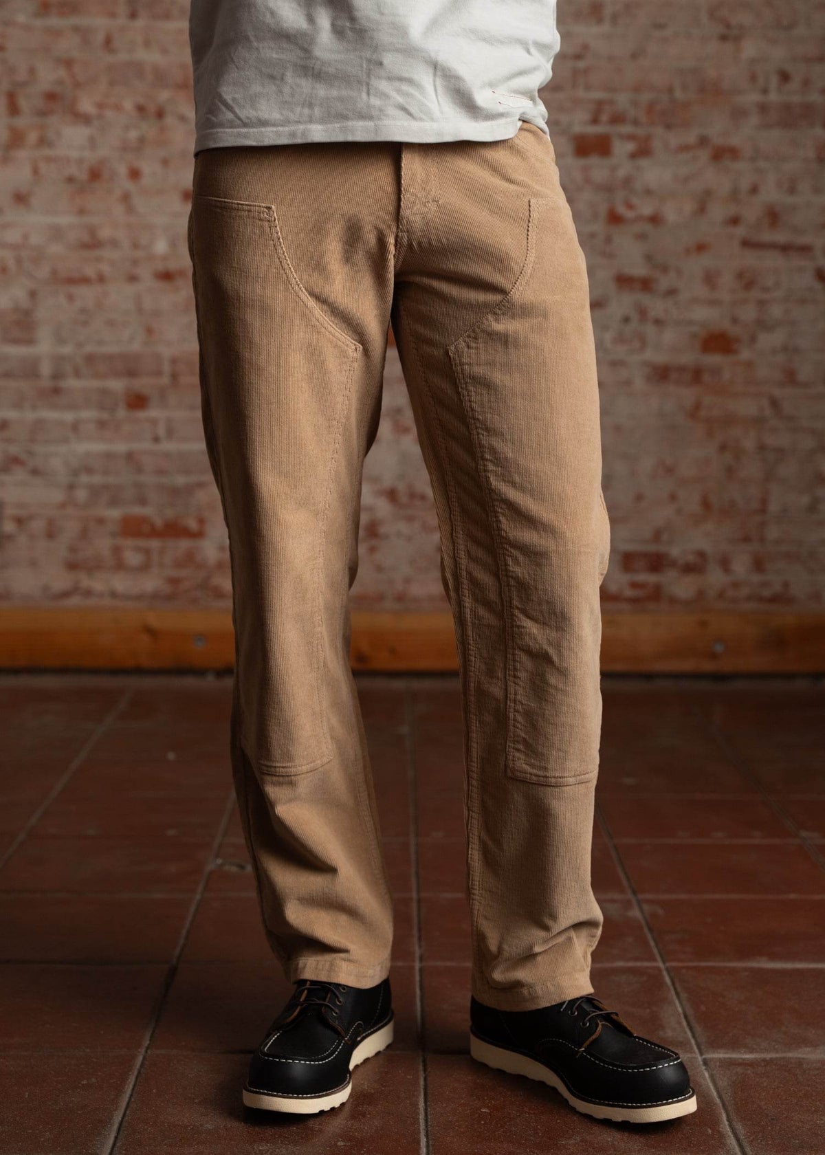 Man in beige pants and black shoes stands on a tiled floor against a brick wall, wearing a white shirt.