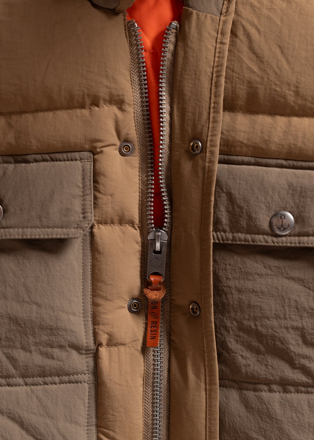 Close-up of a brown jacket with a visible metal zipper and orange lining, featuring a leather pull tab labeled "Iron and Resin."