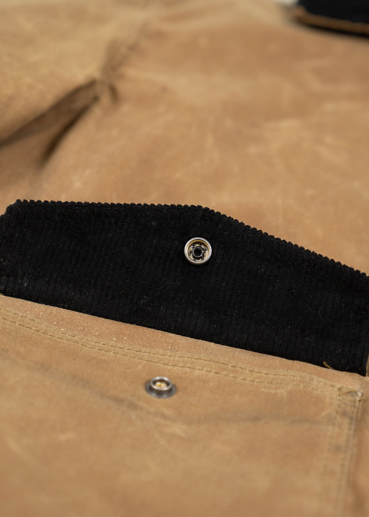 Close-up of a brown jacket pocket with a black corduroy flap and metal snap button, highlighting texture and detail.