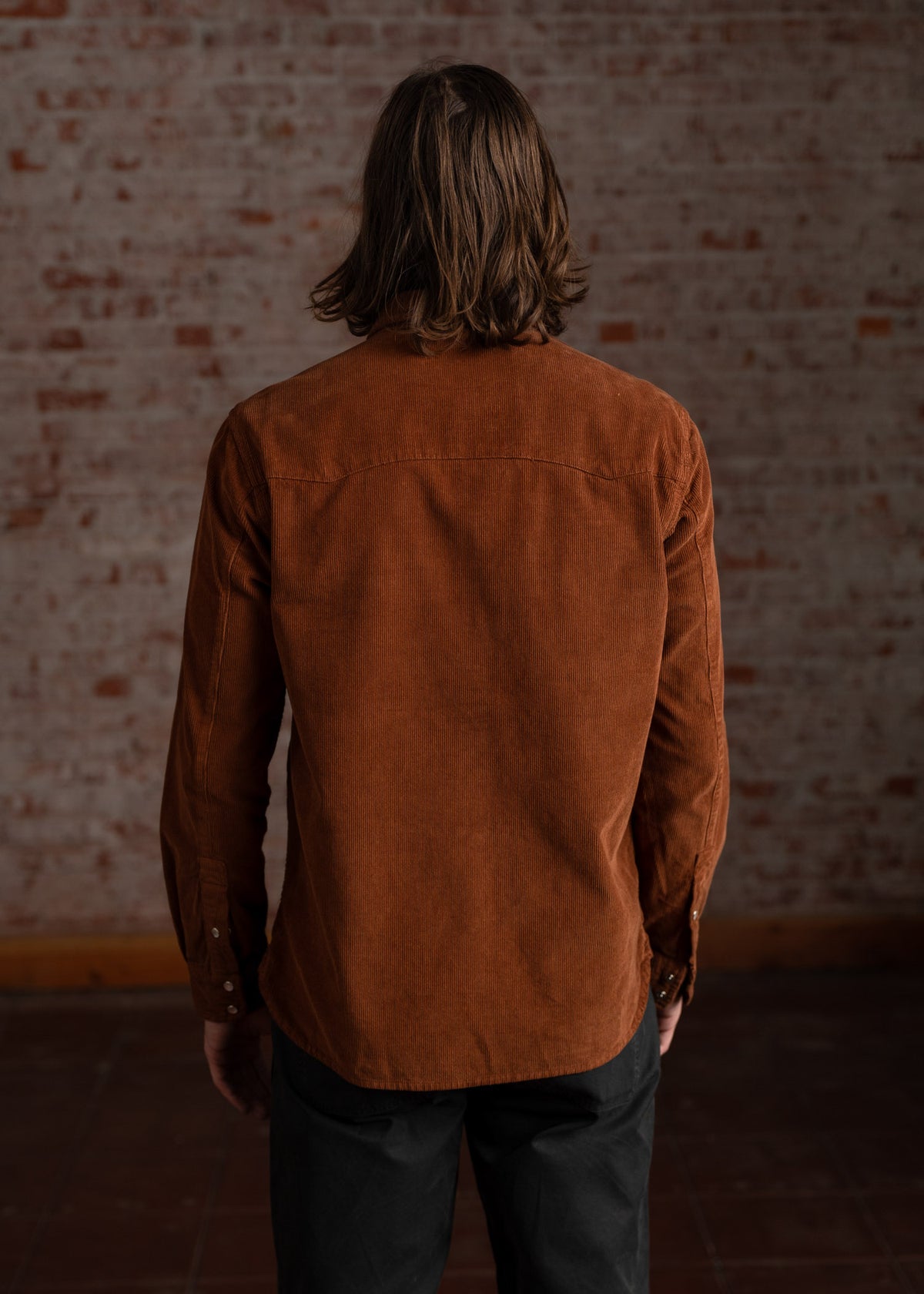 A person with long hair stands facing a brick wall, wearing a brown corduroy shirt and black pants, in a dimly lit room.