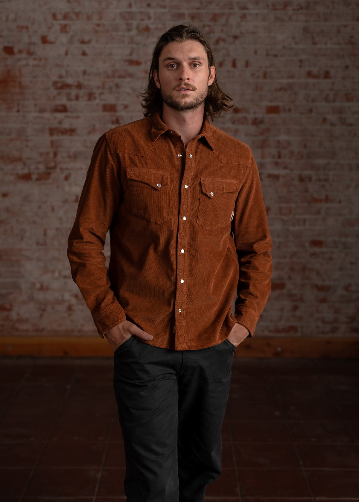 Man in a brown corduroy shirt and black pants stands confidently against a rustic brick wall, hands in pockets.