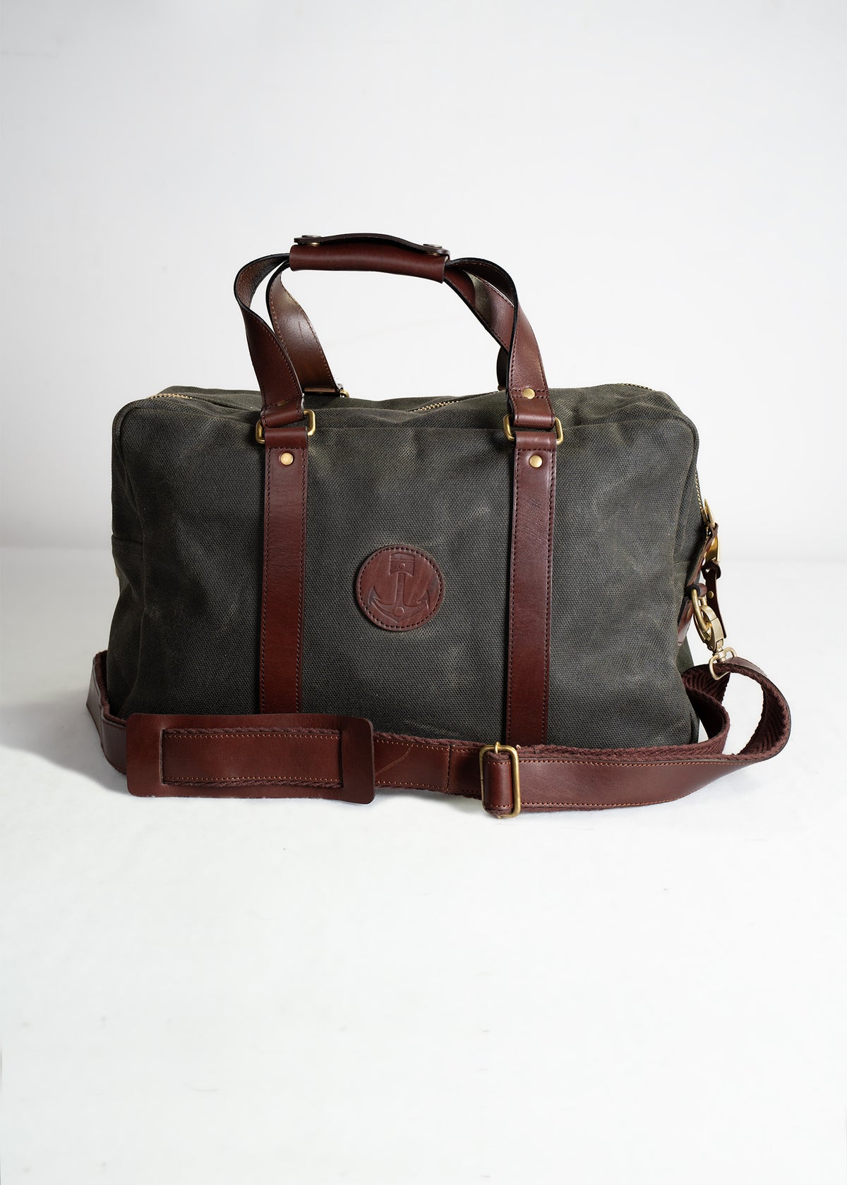 A stylish canvas and leather duffle bag with a shoulder strap, featuring a round embossed logo, sits on a plain white surface.