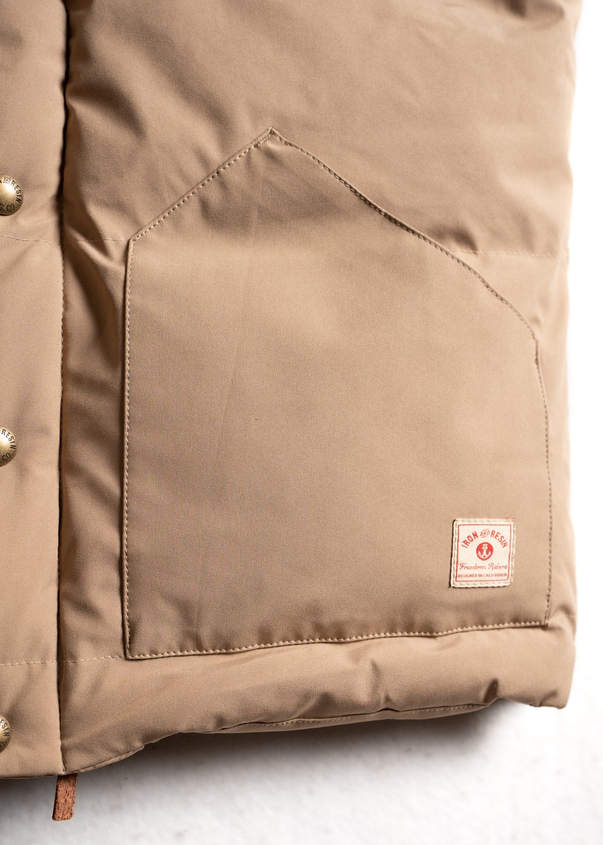 Close-up of a tan jacket pocket with a red "Rock & Resc" label, featuring metal buttons on the left side, against a light background.