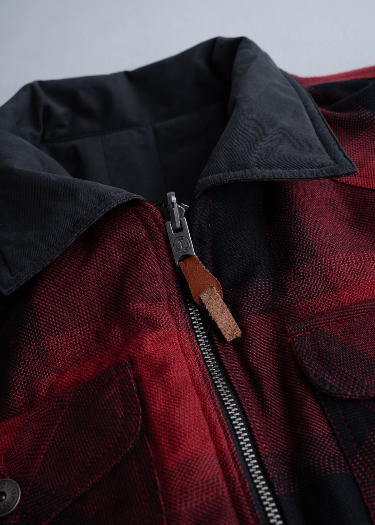 Close-up of a red plaid jacket with a black collar and a metal zipper featuring a brown leather pull tab, lying on a flat surface.