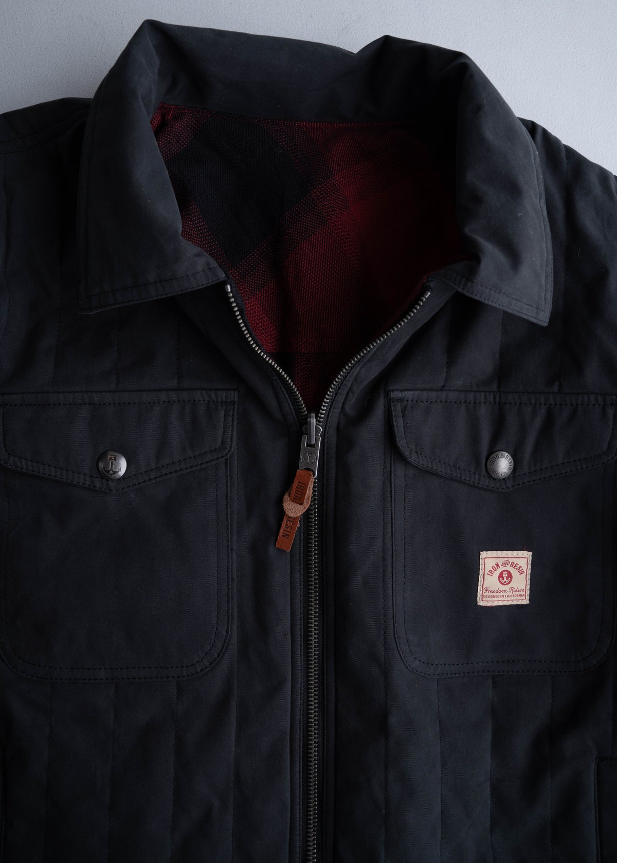 Black jacket with a red plaid lining, featuring a zipper, two front pockets, and a small logo patch on the right pocket.