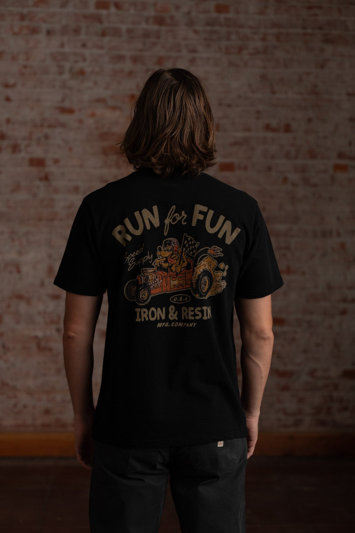 Man in black "Run for Fun" shirt with vintage car design stands against a brick wall, showcasing Iron & Resin branding.