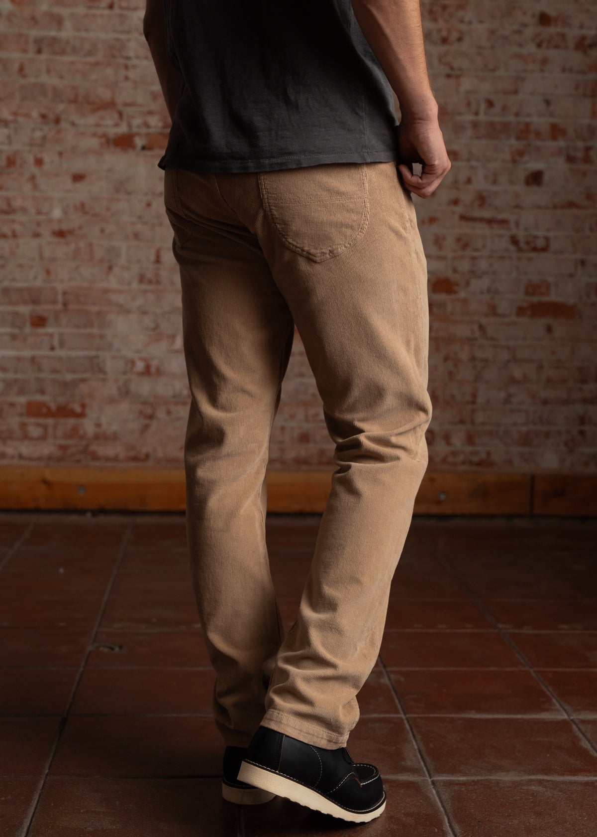 Man in beige pants and black shoes stands on a tiled floor against a brick wall, facing away, with hands by his side.