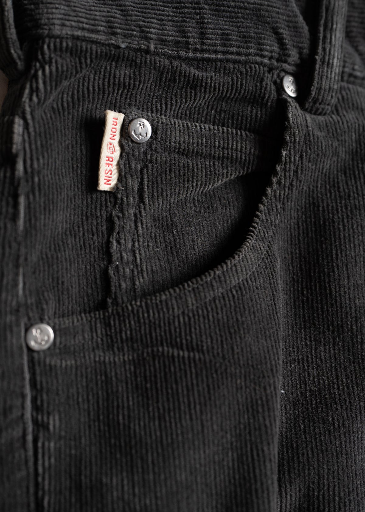 Close-up of black corduroy pants with visible Iron & Resin label and silver rivets, highlighting texture and brand detail.